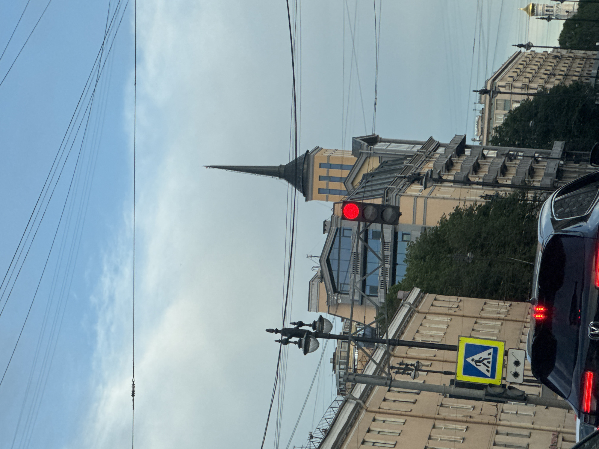 The image depicts a street scene in Saint Petersburg, Russia, likely during daylight with overcast skies. In the foreground, the rear of a black car is visible, displaying illuminated red brake lights. Adjacent to the car, a pedestrian crossing sign is mounted on a pole, featuring a blue square with a white border and a white pictogram of a pedestrian walking. Below this sign, another smaller sign displays a white pictogram of eyeglasses.
Dominating the mid-ground are several buildings and urban infrastructure. A traffic light shows a red signal. A distinctive building with a tall, pointed spire is visible, rendered in yellow and blue. A street lamp with two globes stands prominently to the left. Various electrical wires crisscross the sky above the street. The buildings along the street are of varying architectural styles, with balconies and windows evident. Lush green trees are interspersed between the structures.
In the background, more buildings line the street, and a golden dome, characteristic of Orthodox churches, can be seen in the distance, suggesting the presence of a significant religious structure. The overall impression is a busy urban environment in Saint Petersburg.