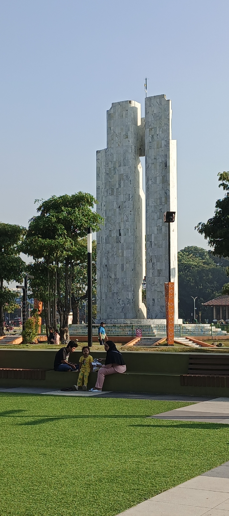 A vertical, eye-level shot captures a public park in Sidoarjo, Indonesia, with a prominent, modern marble monument as the central focus. The monument, composed of two tall, narrow rectangular pillars with a gap between them, is clad in polished marble tiles exhibiting a variegated grey and white pattern. A small flag, yellow and white, flies from the top of the left pillar. In the foreground, a family of three, two adults and a child, are seated on a dark green bench. A young child, wearing yellow pajamas, sits in the middle, facing the camera. To the child's right is an adult in pink pants and a black headscarf, and to the child's left is an adult in a black t-shirt. They appear to be conversing or engaged in quiet activity, with the adult in pink holding a small item. Behind them, the park features lush green grass, manicured trees, and walkways. Further in the background, other individuals are visible, some walking and others seated, suggesting a leisurely day in the park. The sky is clear and bright blue, indicating fair weather.