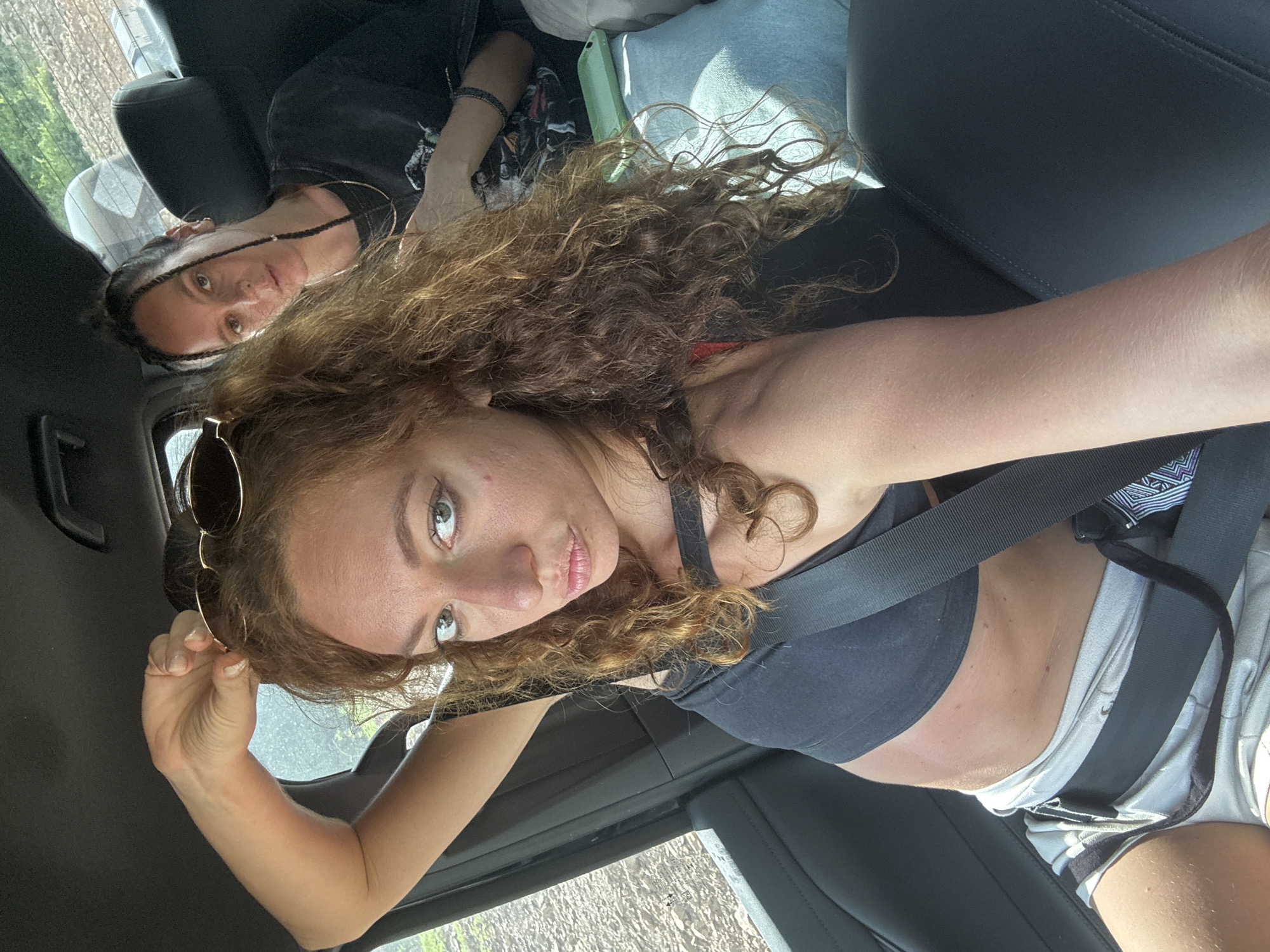 In the image, two individuals are seated inside a vehicle, possibly an SUV, which appears to be in motion as suggested by the blurred outdoor scenery visible through the window. The person in the foreground, a woman with curly brown hair, is taking a selfie. She is wearing a black sports bra and light-colored shorts, with her seatbelt fastened. Her left arm is raised, and she is looking towards the camera with her mouth slightly pursed. Behind her, another woman with dark hair styled in braids is visible, looking forward with a neutral expression. The interior of the car is dark, with black seats and headrests. A light blue pillow and a green phone case are on the seat behind the women. The outdoor environment appears to be rural, with dry, uneven ground visible. The exact location within Tân Phong, Vietnam cannot be determined from the visual information provided.