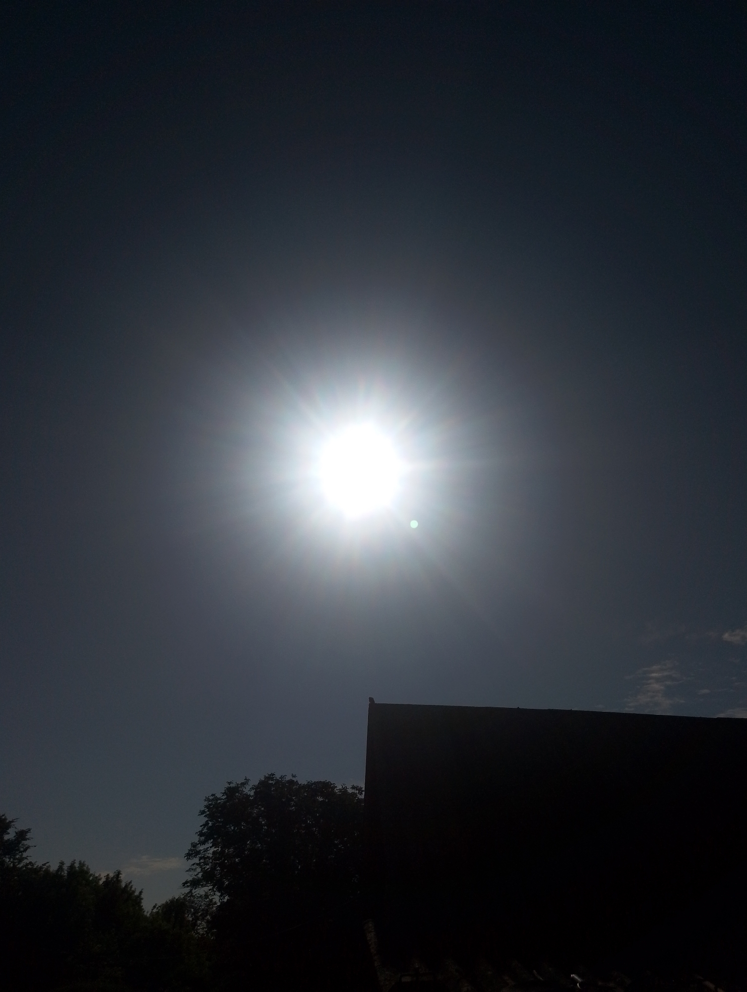 The image shows a bright sun in a dark sky, with the sun's rays radiating outward. In the foreground, the silhouettes of trees and a dark, angled roof are visible against the bright sky. There are no people or specific landmarks of Ustyvytsia, Ukraine identifiable in the image. The overall scene depicts a sunny day with a strong sun.