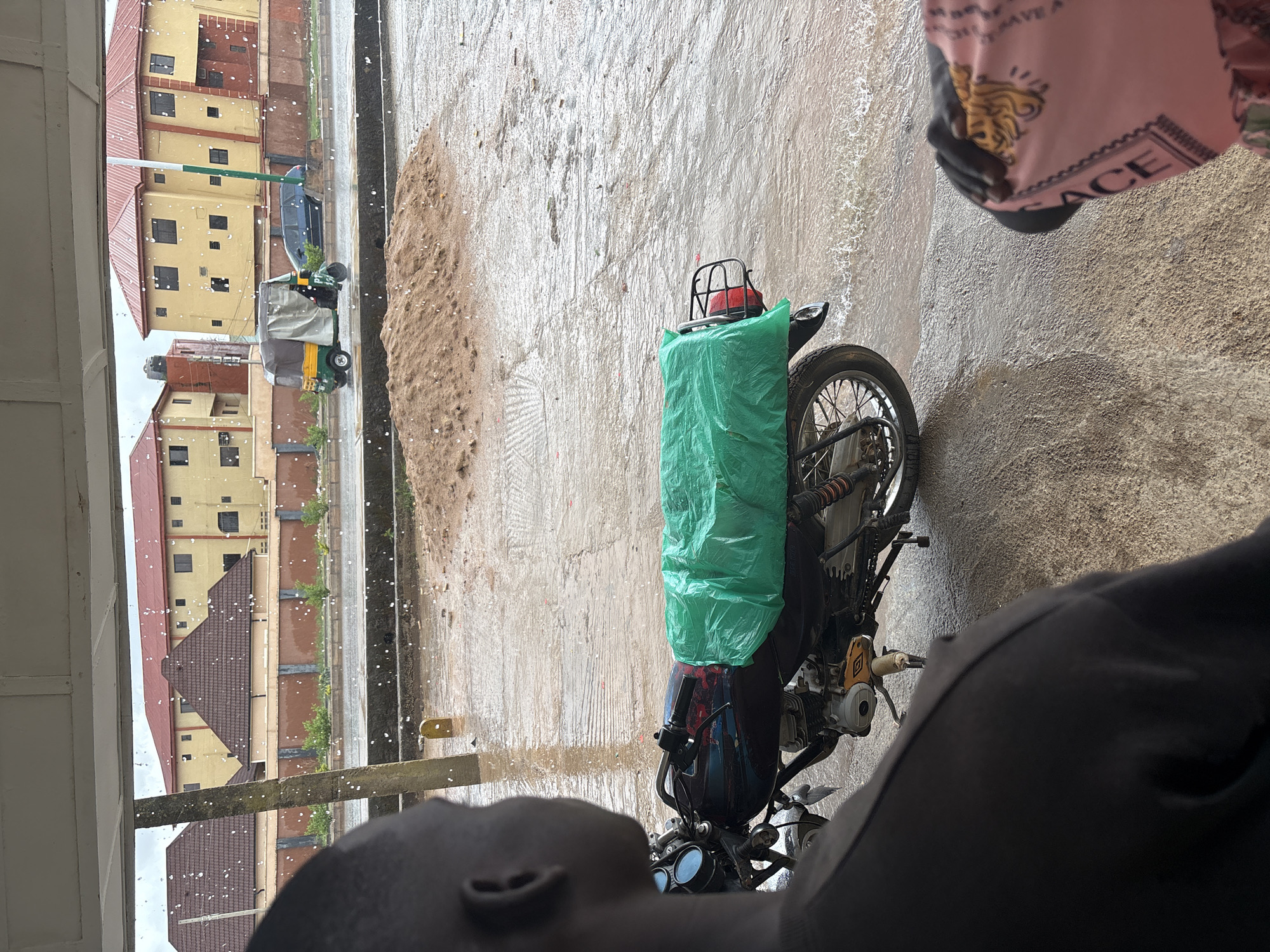 A motorcycle is parked on a paved surface next to a raised concrete area. Raindrops are visible. A three-wheeled vehicle is parked on the street nearby. Buildings are visible in the background. The image appears to be taken from under a shelter. A person's face is visible in the foreground. A person is visible wearing a pink shirt in the foreground. The location is likely Bukuru, Nigeria.