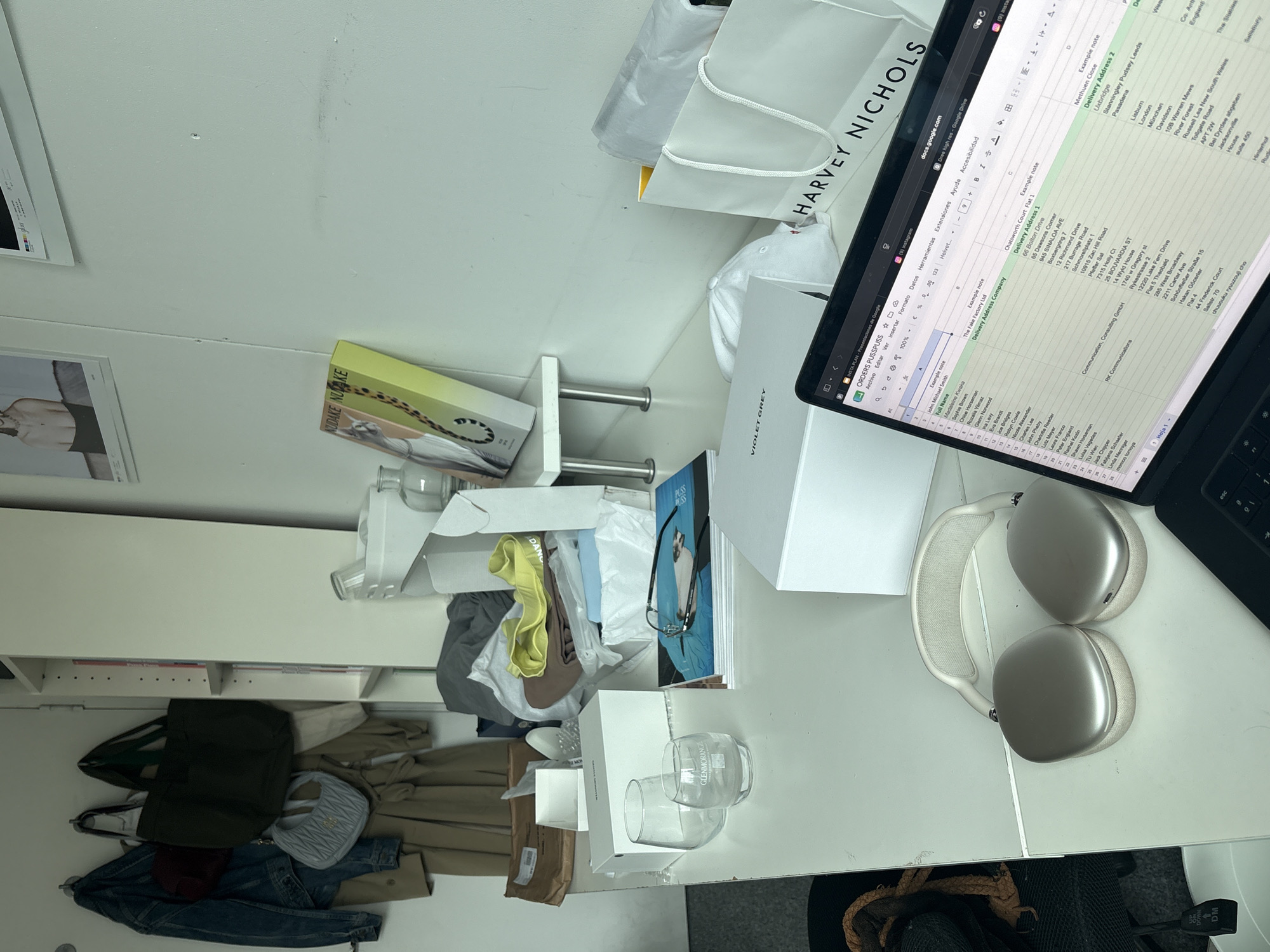 An overhead view captures a busy, white desk or workspace filled with various personal items and work-related materials. A silver laptop, open to a Google Sheets spreadsheet, dominates the upper right portion of the desk. To its left are a pair of silver Apple AirPods Max headphones.
The desk surface is cluttered with several white shopping bags, one prominently featuring "HARVEY NICHOLS". A white box labeled "VIOLET GREY" is also present, alongside two clear glass tumblers, one with "GLENMORANGIE" etched on it. A yellow-green box with the text "NUDAKE NUAKE" stands upright on a shelf-like structure that protrudes from the wall. Various clothes, including a denim jacket, a trench coat, a dark green tote bag, a light blue quilted purse, and a white baseball cap, are draped or folded on and around the desk, suggesting either recent unpacking or a casual storage area. Below the desk, a black office chair is partially visible.
The laptop screen displays a spreadsheet titled "ORDERS PUSSPUSS" from "docs.google.com", showing columns for "Full Name", "Delivery Address Company", "Delivery Address 1", and "Delivery Address 2". Visible names include "John Michael Smith" and "Madaleine Kwaka", with addresses mentioning locations like "London", "England", and "Communication, Consulting GmbH". A framed image on the white wall to the left has "ghos" visible. The overall scene suggests an active personal workspace, possibly a home office, with items being organized or recently used.