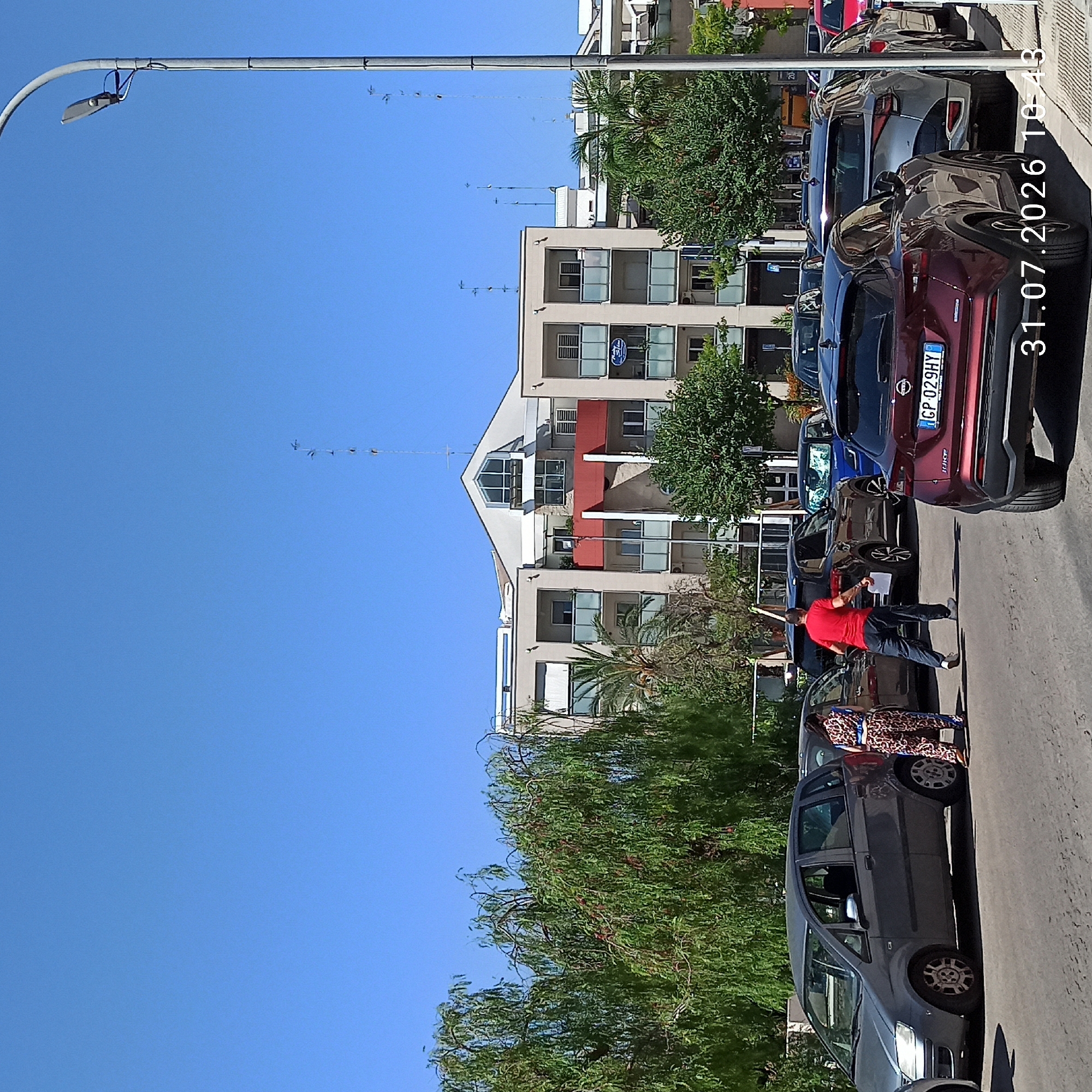 The image depicts a street scene in Modugno, Italy, on July 31, 2026, at 10:43 AM. The sky is clear and blue. In the foreground, a maroon Nissan Juke is parked, with its license plate "CP 029HY" visible. To its left, a silver SUV is parked, followed by a dark SUV and then a blue hatchback. Further down the street, a gray MPV is parked. A woman wearing a leopard-print outfit with a blue belt is standing next to the gray MPV. A man in a red t-shirt and dark pants is walking towards the dark SUV, holding a white piece of paper. Several trees with green foliage line the street, and a modern building with balconies and windows is visible in the background. A lamppost stands on the right side of the image.