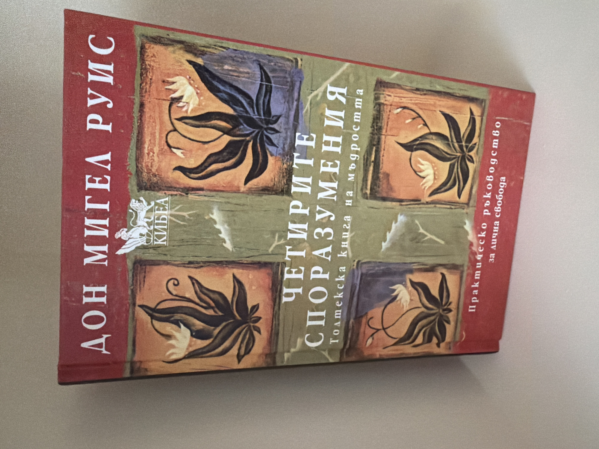 The image shows the cover of a book titled "The Four Agreements" by Don Miguel Ruiz. The book is displayed from a slightly elevated, angled perspective, with the spine on the left and the front cover facing the viewer. The background is a plain, light-colored surface, likely a table or shelf. The book cover features a red border and a central design with four square panels, each containing an illustration of a plant with stylized flowers. The text on the cover is in white and appears to be in Cyrillic script. The publisher's logo, a lion, is visible near the top center of the cover. No people are visible in the image. The book appears to be in good condition.
Based on the information provided, it is not possible to determine the location in the city of Pazardzhik, Bulgaria. The image only shows the book cover.