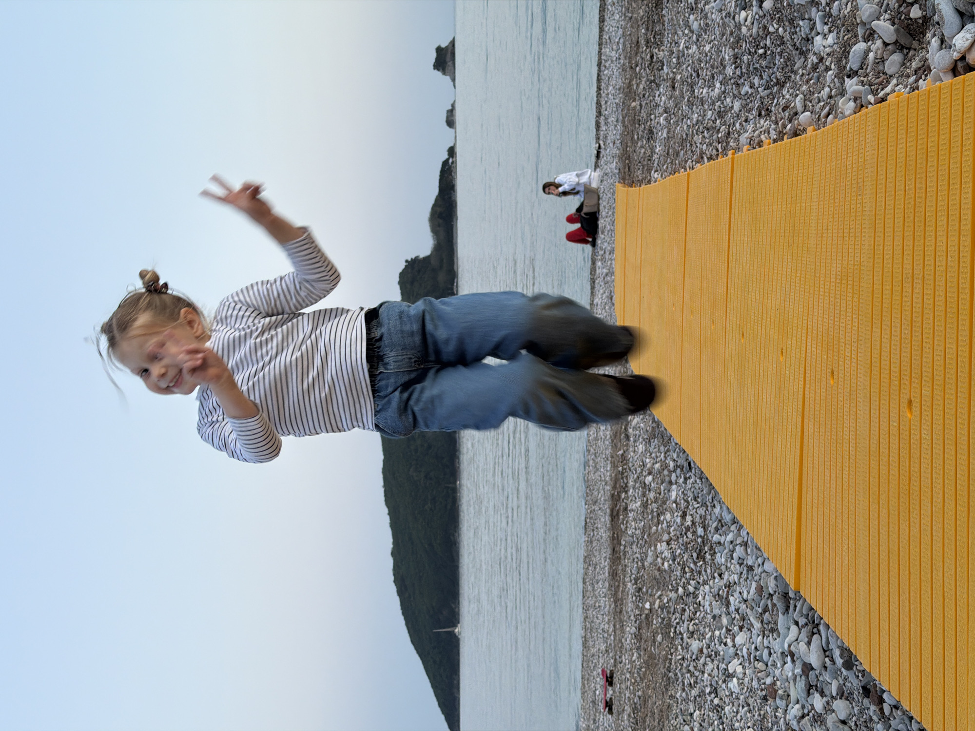 A young girl wearing a striped shirt and blue jeans is captured mid-air, giving a peace sign with both hands. She is suspended against a backdrop of the sky, a body of water, and an island with dense vegetation. In the distance, two people are seated on the beach, next to a yellow walkway leading onto the beach. The beach is covered in small, grey stones. Based on the elements in the image, the location is likely a beach in or near the city of Budva, Montenegro.