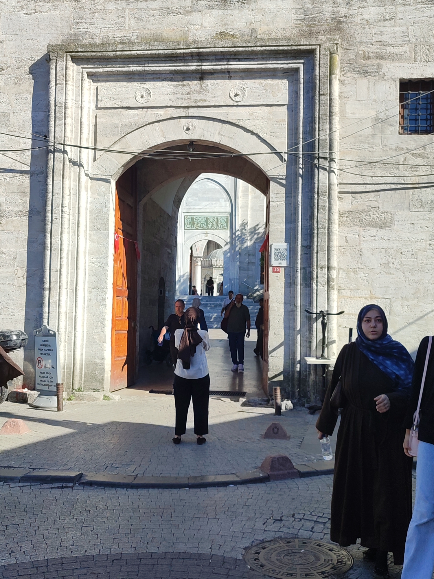 The image shows a view looking through a large stone archway, possibly an entrance to a courtyard or a religious building. The archway is made of light-colored stone with intricate carvings. Sunlight casts strong shadows across the scene.
In the foreground, to the left, is a sign with Turkish text and a red circle with a diagonal line, indicating prohibition. There is also a QR code displayed on a small sign. A wooden door is ajar to the right of the archway.
Several people are visible walking through or standing near the archway. In the immediate foreground, on the right, a woman wearing a dark abaya and a blue hijab is holding a plastic water bottle. She is looking towards the left. Further back, a woman in a white shirt and black pants with a brown hijab is walking away from the camera. Other figures are seen in the background, some walking, some partially obscured.
Beyond the archway, another arched structure is visible, with a dome and intricate patterns on its facade. The ground is paved with cobblestones.
The scene appears to be a public space, possibly the grounds of a mosque or historical complex, given the architectural style. The location is Üsküdar, Turkey.