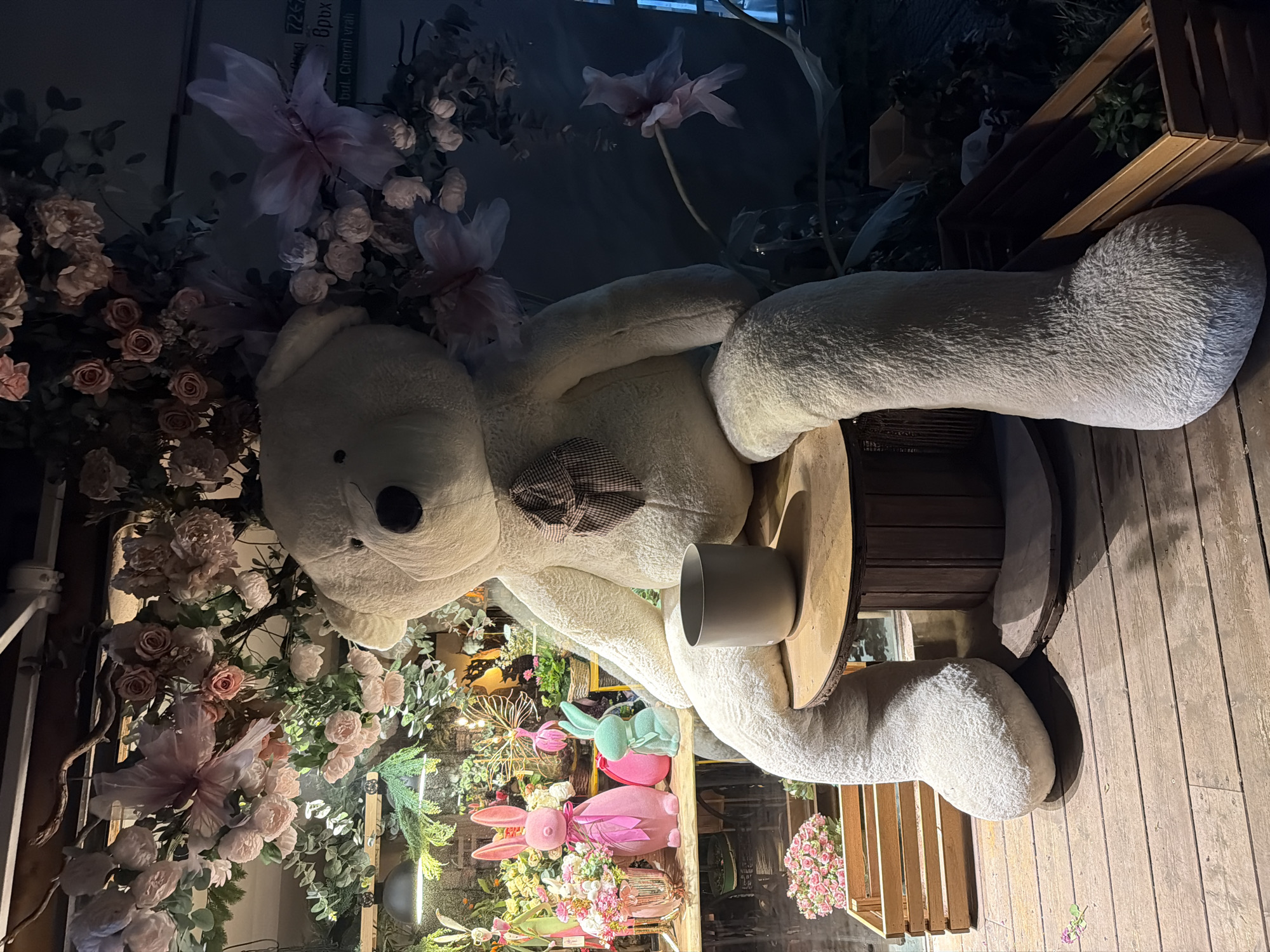 A large, cream-colored teddy bear sits on a wooden platform inside a shop or cafe setting, in Sofia, Bulgaria. The bear is positioned facing left, with a brown and white checkered bow tie. The bear is holding a gray cup on a wooden table. Various floral arrangements, including roses and decorative butterflies, surround the bear. Below, there are pink bunny figurines and other decorations. The wooden platform sits on a wooden floor. Overhead, there's a glimpse of a window and floral arrangements continue into the darker areas of the interior.