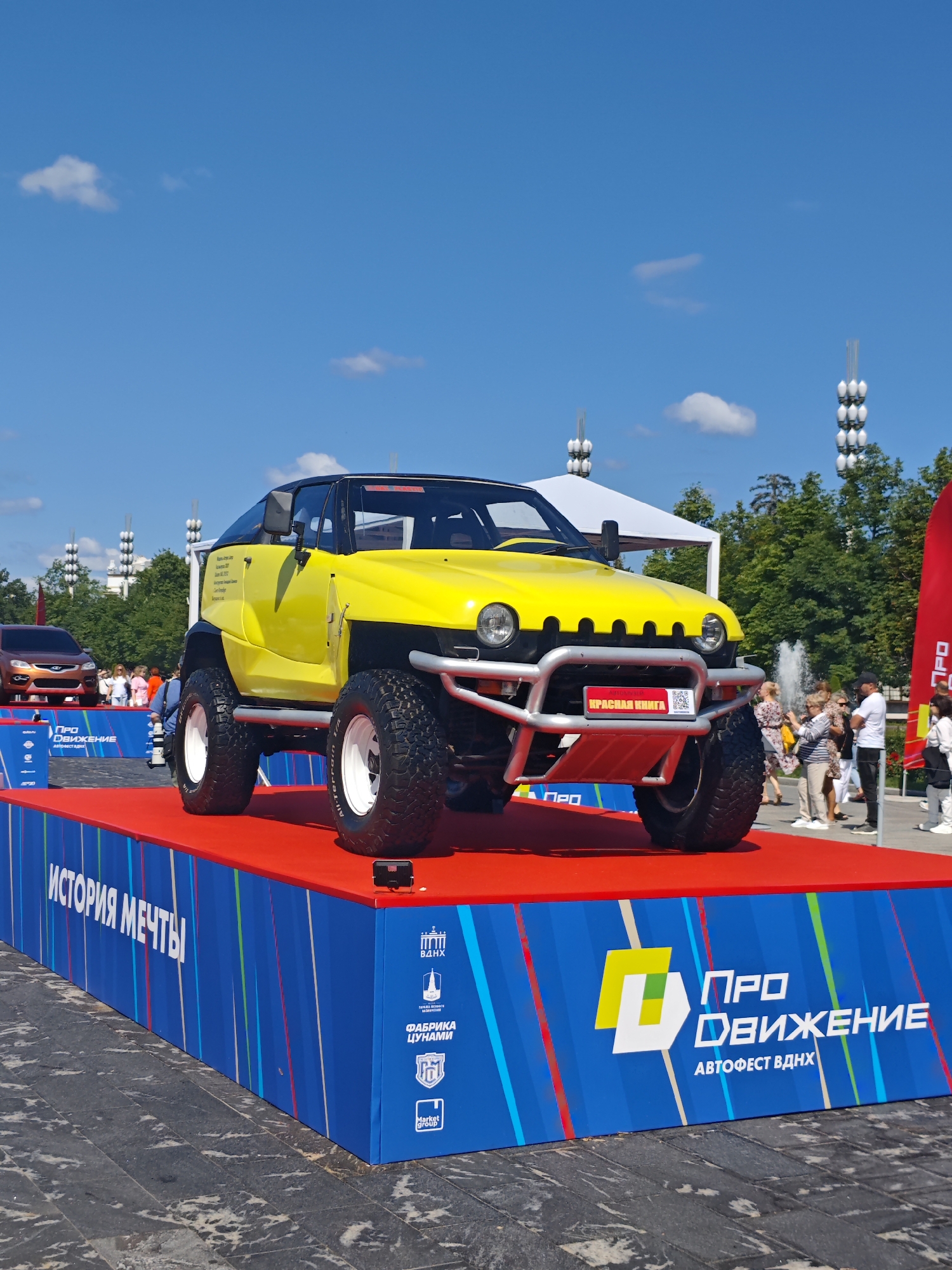 A bright yellow, off-road vehicle with large tires is prominently displayed on a red platform in an outdoor urban setting. The vehicle has a black convertible top and a silver bumper. In the background, people are visible walking and standing near trees and fountains. The scene appears to be an event or exhibition, suggested by the blue and red checkered banners with text in Russian, including "ИСТОРИЯ МЕЧТЫ" (History of Dreams) and "ПРО ДВИЖЕНИЕ АВТОФЕСТ ВДНХ" (PRO Movement Autofest VDNKh). This indicates the event is taking place at VDNKh, a large exhibition center in Moscow, Russia, known for its pavilions and fountains. The sky is clear and blue with a few scattered white clouds. The pavement in the foreground has a speckled, worn appearance.
