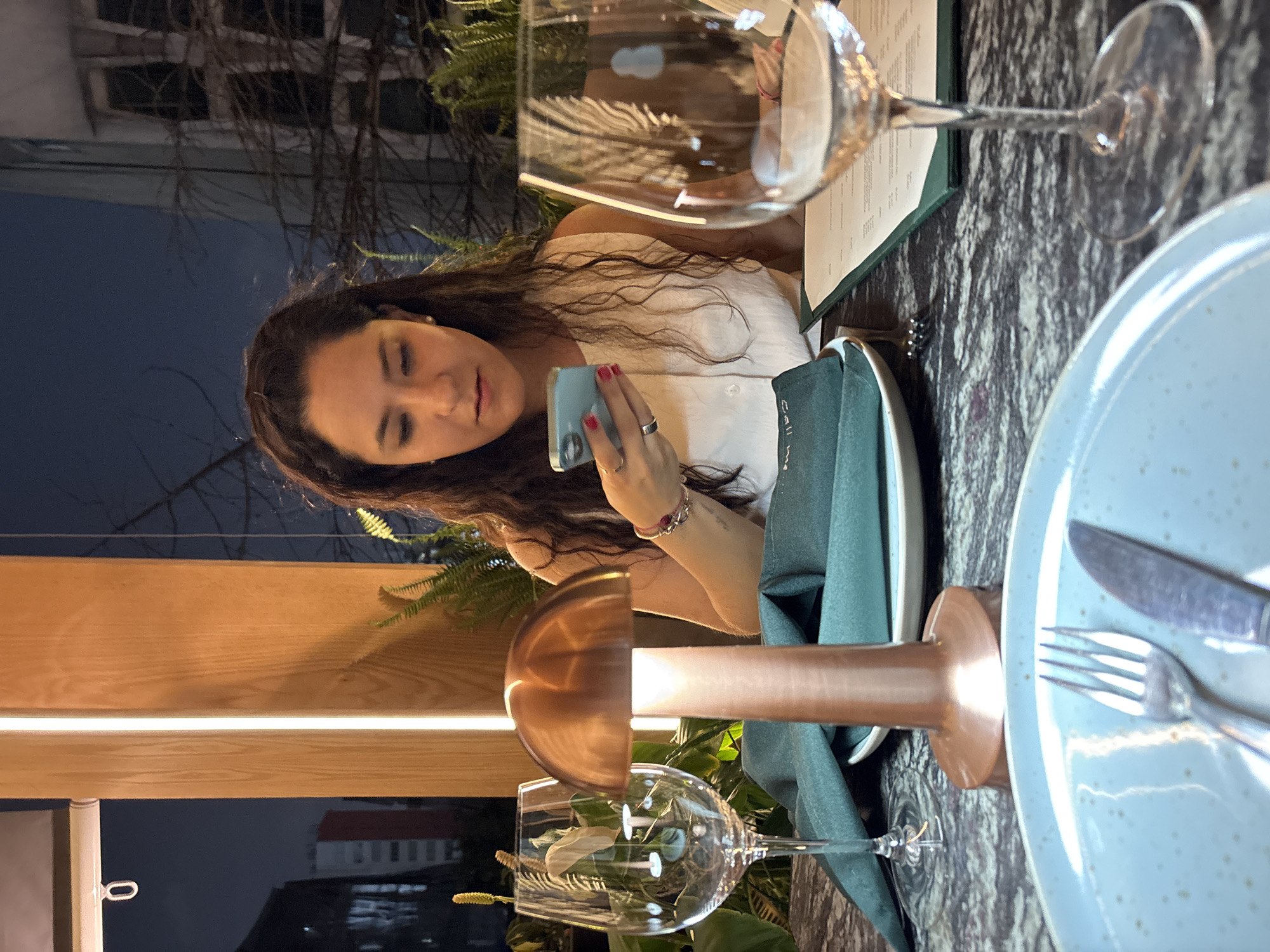 A medium shot depicts a woman seated at a restaurant table in Mexico City, Mexico. She has long, curly brown hair and wears a white top. She is looking at a blue smartphone in her hands. Her left hand has a ring on one finger.
The table is set with a light blue plate, a fork, a knife, a green napkin, a menu, and two wine glasses. The tabletop has a gray, marbled pattern. A copper-colored lamp illuminates the setting.
Outside the restaurant, there is a view of buildings, suggesting the scene is indoors but near an outdoor environment. Fern-like plants are visible near the woman.