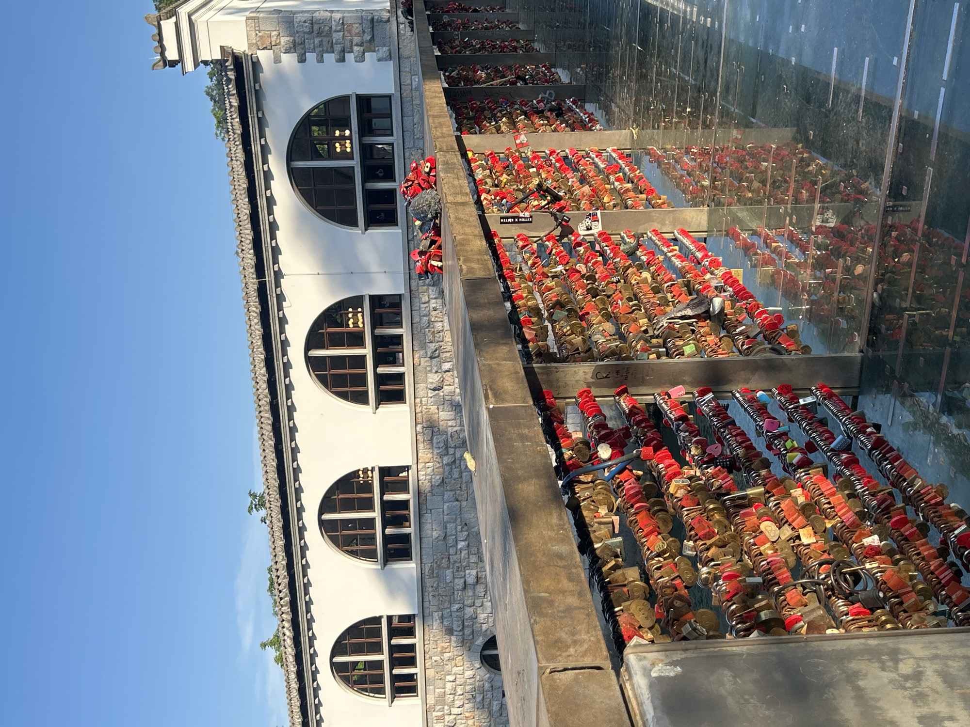 This is a vertically oriented, eye-level, medium shot of a bridge adorned with countless love locks. The scene is bathed in bright daylight, suggesting a clear day. The bridge's structure is visible, with a white, stone-accented building on the left featuring arched windows and a decorative edge. To the right, a railing made of glass and metal separates the bridge walkway from the locks, which are densely packed in rows. The locks are predominantly red and gold, with some in other colors like pink and blue, and various shapes and sizes. Some appear new, while others show signs of weathering. The sheer volume of locks implies a long-standing tradition. A few individuals are visible on the bridge, one standing near the locks, possibly adding a new one, and another further down. The setting appears to be a popular tourist spot, given the nature of the display. The overall impression is one of romance and commitment, with the sunlight highlighting the vibrant colors of the locks. Some text is visible on a few locks, though it is too small and indistinct to be read.