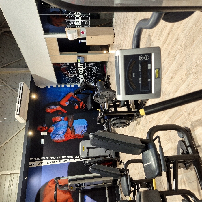 The image displays the interior of a Keepcool Social Sports Club gym located in Strasbourg, France. The facility is unoccupied, featuring a light wooden floor and a high ceiling with visible structural elements and a suspended white HVAC unit. Various Technogym exercise machines are arranged throughout the space.
In the foreground, a gray digital display unit from a Technogym cardio machine is visible, showing readouts for 'ROM', 'REPS', and 'WEIGHT'. The mid-ground contains multiple black and silver Technogym strength training machines with yellow adjustment points, including several seated units and a plate-loaded machine with black weight plates.
The rear wall is adorned with a large mural depicting a man in a blue shirt and a woman in a black sports top, with the gym's name, 'SOCIAL SPORTS CLUB / KEEPCOOL', printed vertically. To the right, a glass-partitioned section with white text patterns, including 'WORKOUT', reveals a wooden cabinet supporting a wall-mounted paper towel dispenser and a hand sanitizer pump bottle. A small digital screen on the wall also displays 'WORKOUT'.