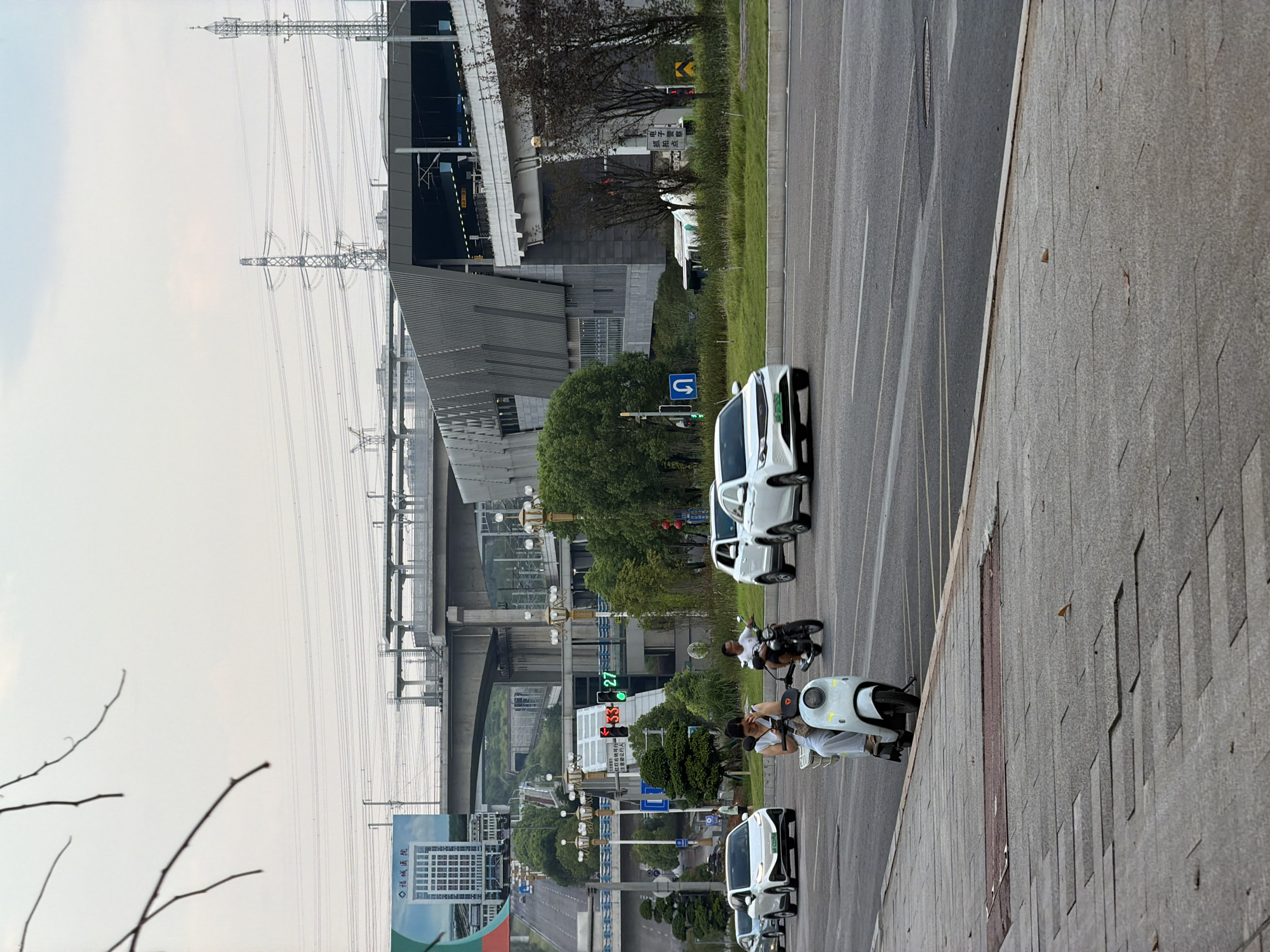A daytime, eye-level shot captures a busy street scene in Jiangjin, China, characterized by a multi-layered urban environment with evident infrastructure development. In the foreground, a grey concrete sidewalk with visible paving stones and scattered leaves runs along the bottom edge. The main focus of the street is a two-lane asphalt road where several vehicles are in motion. A white electric scooter with a rider wearing light-colored clothing is prominently positioned on the right lane, seemingly at a standstill or moving slowly. To its right, a white motorcycle carries a rider wearing a white shirt. Several white cars are also present, including one in the left lane turning right and another in the right lane moving forward.
The mid-ground reveals a complex network of elevated structures. A concrete overpass, likely for a highway or an elevated railway, dominates the scene, with its supporting pillars and bridge deck. Beneath this, a modern, multi-story building with a distinctive patterned facade, possibly a transportation hub or commercial complex, is visible. Traffic lights are also present, with a red light illuminated on one signal and a green light on another displaying the number 27. Trees and green spaces are interspersed between the structures, suggesting a planned urban landscape.
In the background, the sky is overcast with a soft, diffused light. Electrical power lines and transmission towers are clearly visible, indicating the presence of significant power infrastructure. A large billboard, partially obscured by branches in the upper left, displays text and an image of a tall building. The overall impression is one of a rapidly developing city with advanced transportation and infrastructure.
