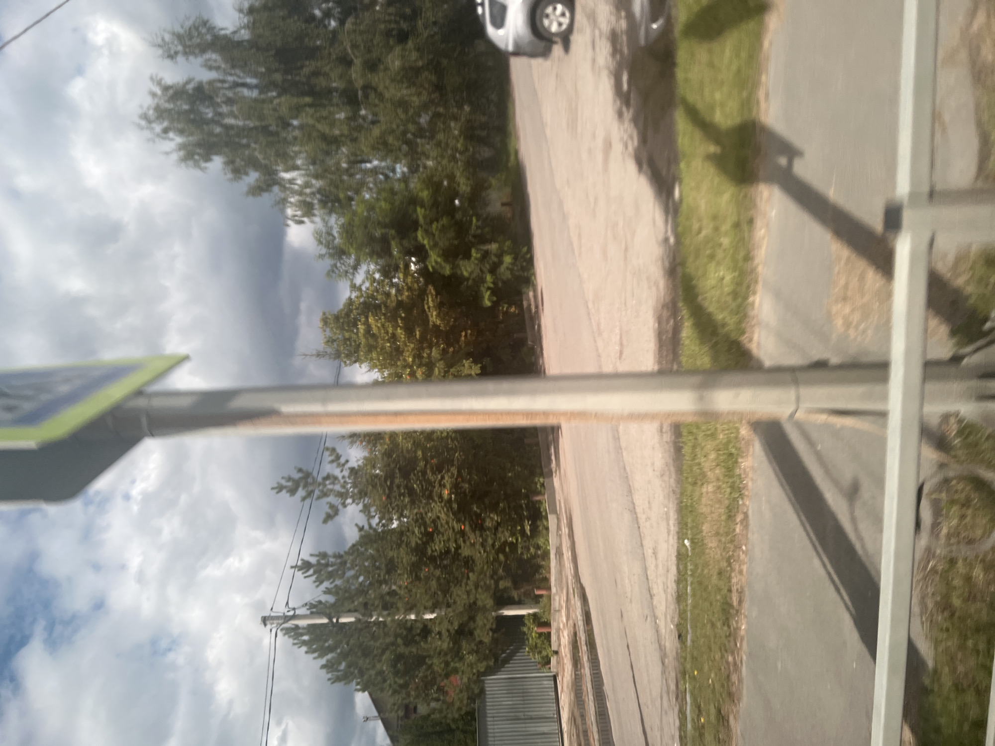 The image depicts a street scene in Ulyanovsk, Russia, likely taken during the daytime. In the foreground, a blurred metallic railing is visible, suggesting the viewpoint is from behind a fence or barrier. A light-colored pole with a rectangular sign, partially obscured and out of focus, rises vertically in the center of the frame. This sign appears to be a traffic or directional marker.
The middle ground shows a paved road with a grass verge. To the left, a corrugated metal fence segment is visible. Beyond the road, dense greenery dominates, with various trees and bushes forming a natural backdrop. Some trees exhibit reddish-orange fruit or berries. A modern, silver SUV is parked on the right side of the road, its rear end and one wheel clearly visible.
The background is characterized by a cloudy sky, with patches of blue peeking through. Power lines stretch across the sky, supported by a utility pole. A building with a dark roof can be seen peeking through the trees on the far left. The overall impression is of a quiet residential or suburban area. The lighting suggests it's either overcast or the sun is low in the sky, casting faint shadows. The image's blurriness suggests motion, possibly taken from a moving vehicle.