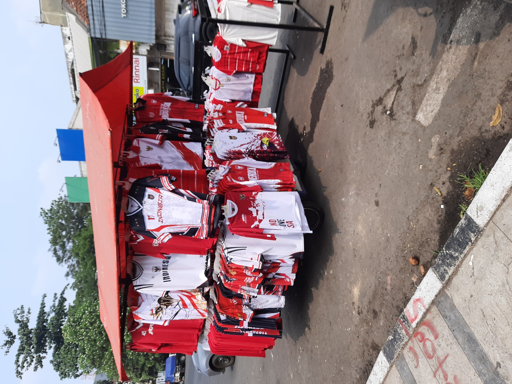 An outdoor market stall displays numerous red and white t-shirts, likely merchandise related to Indonesian sports teams or national pride, in front of a building with a red awning. The stall is set up on a paved street, with a curb and sidewalk in the foreground. A black SUV is parked behind the stall. Trees are visible in the background, along with a Rinnai sign and what appears to be a "TOKO" sign, indicating a shop. The overall scene suggests a commercial street environment in a city.