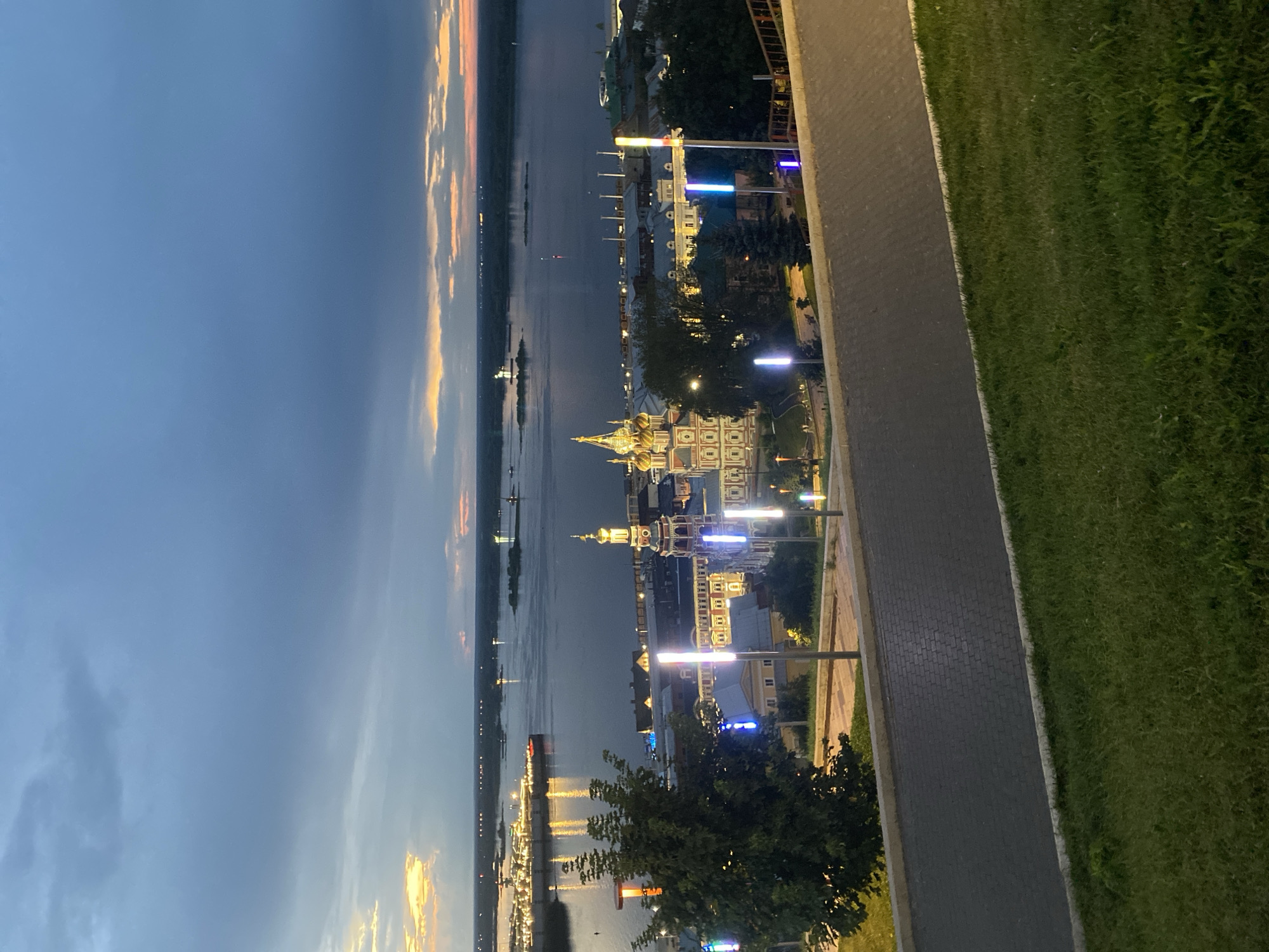 The image captures a panoramic view of the confluence of the Oka and Volga rivers in Nizhny Novgorod, Russia, at dusk. The sky is a dramatic mix of deep blues, grays, and hints of orange and yellow from the setting sun peeking through the clouds.
In the foreground, a paved walkway with cobblestone accents curves towards the right, bordered by a grassy slope. Modern streetlights with vertical blue and white lights line the walkway, illuminating the path and casting colorful reflections.
Further into the scene, a cluster of buildings with ornate architecture, including a prominent church with golden domes and colorful facades, stands on the riverbank. These buildings are illuminated by interior and exterior lights, creating a warm glow. A large body of water, the river, stretches across the midground, reflecting the sky and the lights of the city. On the river, a large, illuminated ship, possibly a cruise liner, is docked or moving, its lights casting a bright reflection on the water. Distant shores are visible, lined with trees and dotted with lights from what appears to be a small town or settlement. Several other smaller islands or landmasses are scattered in the river. The overall atmosphere is serene and picturesque, showcasing the beauty of Nizhny Novgorod at twilight.