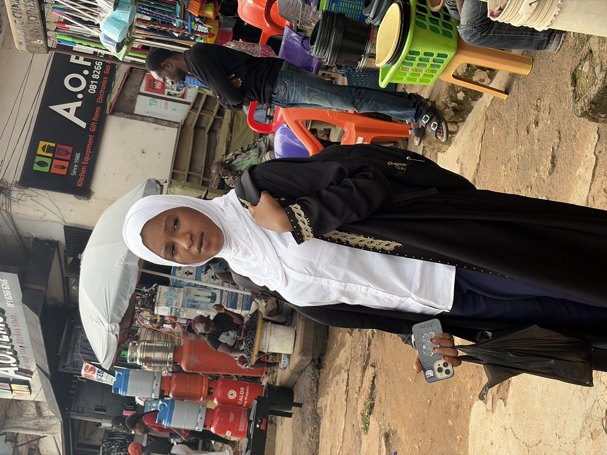 The image captures a street scene in Jos, Nigeria, likely a bustling market area. In the foreground, a woman wearing a white hijab and a black abaya stands holding a black plastic bag and a phone. She is looking directly at the camera with a neutral expression. Behind her, various goods are displayed, including stacks of gas cylinders, brooms, and other household items. Several other individuals are visible in the background, some seated and others standing, engaged in their daily activities. The environment appears to be an outdoor market with stalls and shops lining the street, characteristic of an urban setting in Jos. The ground is paved with rough concrete or dirt, and there are several plastic chairs scattered around, suggesting a place where people gather or conduct business. The overall impression is one of everyday commerce and social interaction within the city.