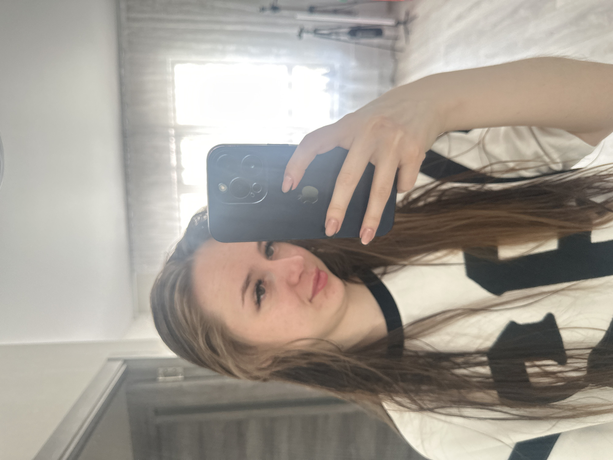 A young woman is depicted taking a mirror selfie in an indoor setting, likely a bedroom or living space. She is shown from the chest up, with long, wavy brown hair framing her face and cascading over her shoulders. She has a light complexion and a neutral to slightly smiling expression, looking directly at the camera.
She holds a dark-colored iPhone with her left hand, partially obscuring her face. The phone's triple camera system and the distinctive Apple logo are clearly visible on its back. Her fingernails are manicured with a light pink polish. She is wearing a white t-shirt adorned with black designs, most notably a clear silhouette of a cat and other abstract black shapes.
The background, which is a reflection in the mirror, features a large, brightly lit window, indicating it is daytime. The natural light casts reflections and highlights across the scene. White bedding or furniture can be seen in the background, and a dark door frame is visible on the left side of the reflection. The overall atmosphere is casual and well-lit.