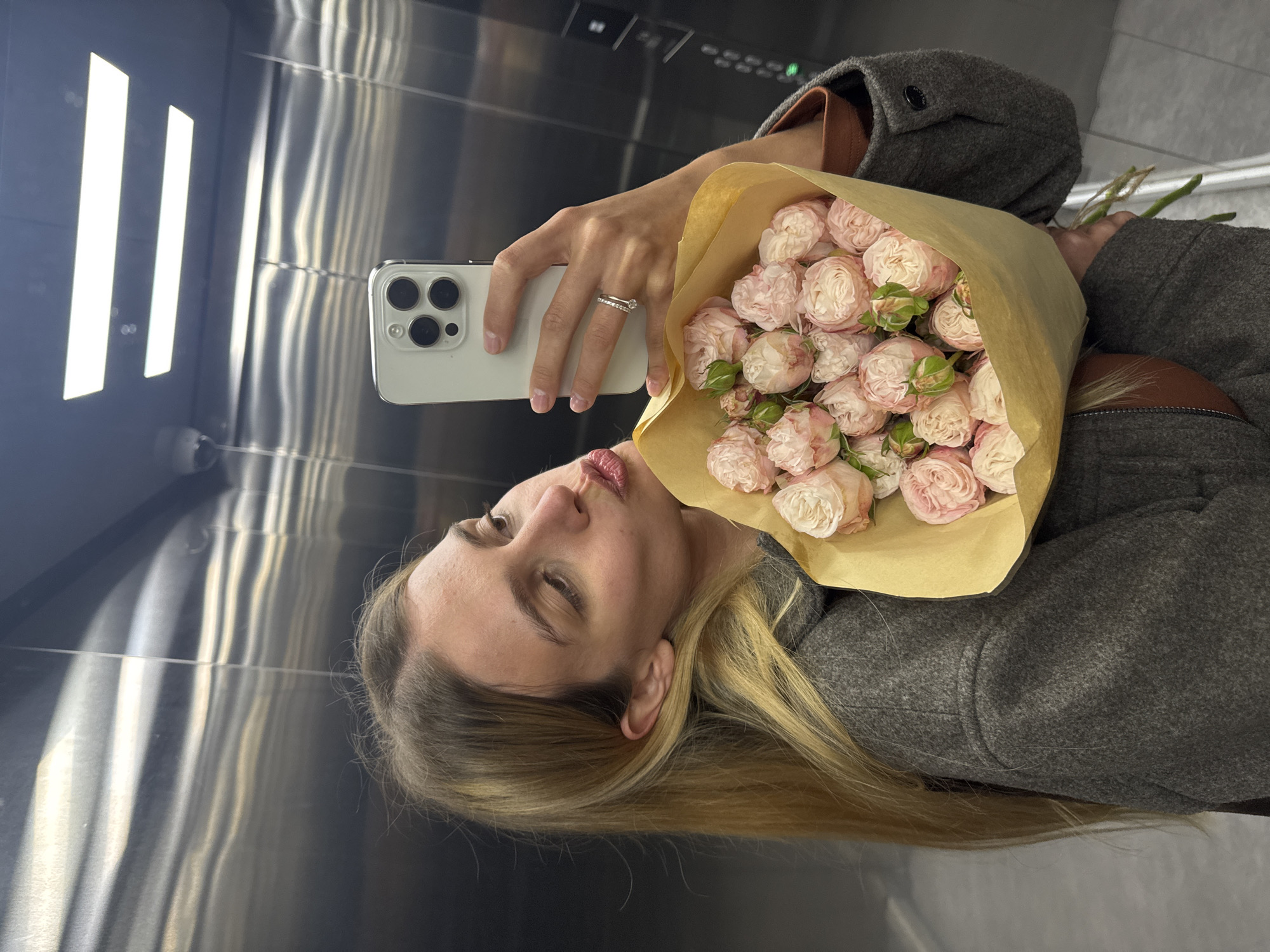A woman is taking a selfie in an elevator. She is holding a bouquet of light pink roses wrapped in brown paper. She is wearing a grey coat and a ring on her finger. Her blonde hair is long and straight. The elevator has stainless steel walls and ceiling with rectangular lighting fixtures. The elevator buttons are visible.