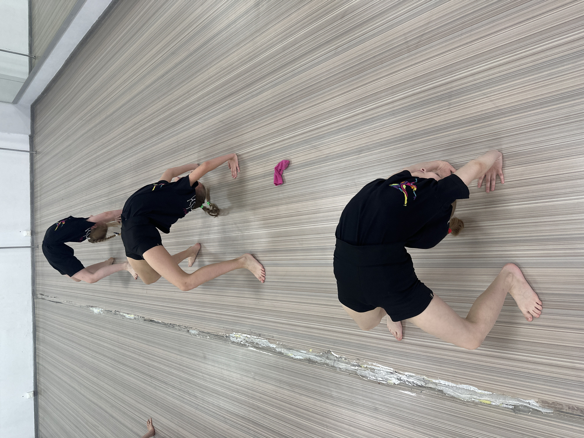 An indoor scene, captured with a rotated perspective, shows two individuals practicing flexibility on a patterned floor in what appears to be a studio in Votkinsk, Russia. Both subjects, likely young women or girls, are seen performing a backbend or bridge-like pose. They are dressed in black t-shirts and black shorts.
The individual on the left is smaller, with light-colored hair tied into pigtails with green hair ties. Her black t-shirt features an abstract, colorful design on the front. Her hands and bare feet are pressed against the floor as she maintains the arch in her back.
The second, larger individual is positioned to the right, also wearing a black t-shirt with a colorful graphic design on the chest and black shorts. She too is in a backbend pose, with her hands and bare feet on the patterned surface.
The floor is a light-colored surface with a distinctive striped pattern running horizontally across the frame. In the middle ground, a long, horizontal strip of damaged or repaired flooring is visible, characterized by a shiny, silver-colored material, possibly tape or putty. A bright pink sock or piece of fabric lies on the striped floor between the two individuals.
On the far left, a large mirror reflects a white wall, a metal railing or bar, and a partial view of what appears to be another person's bare foot near the bottom left corner. The setting suggests a dedicated space for physical training, such as gymnastics or dance. No specific time of day is discernible, and facial expressions are not visible to convey emotion. No clearly legible text is present.