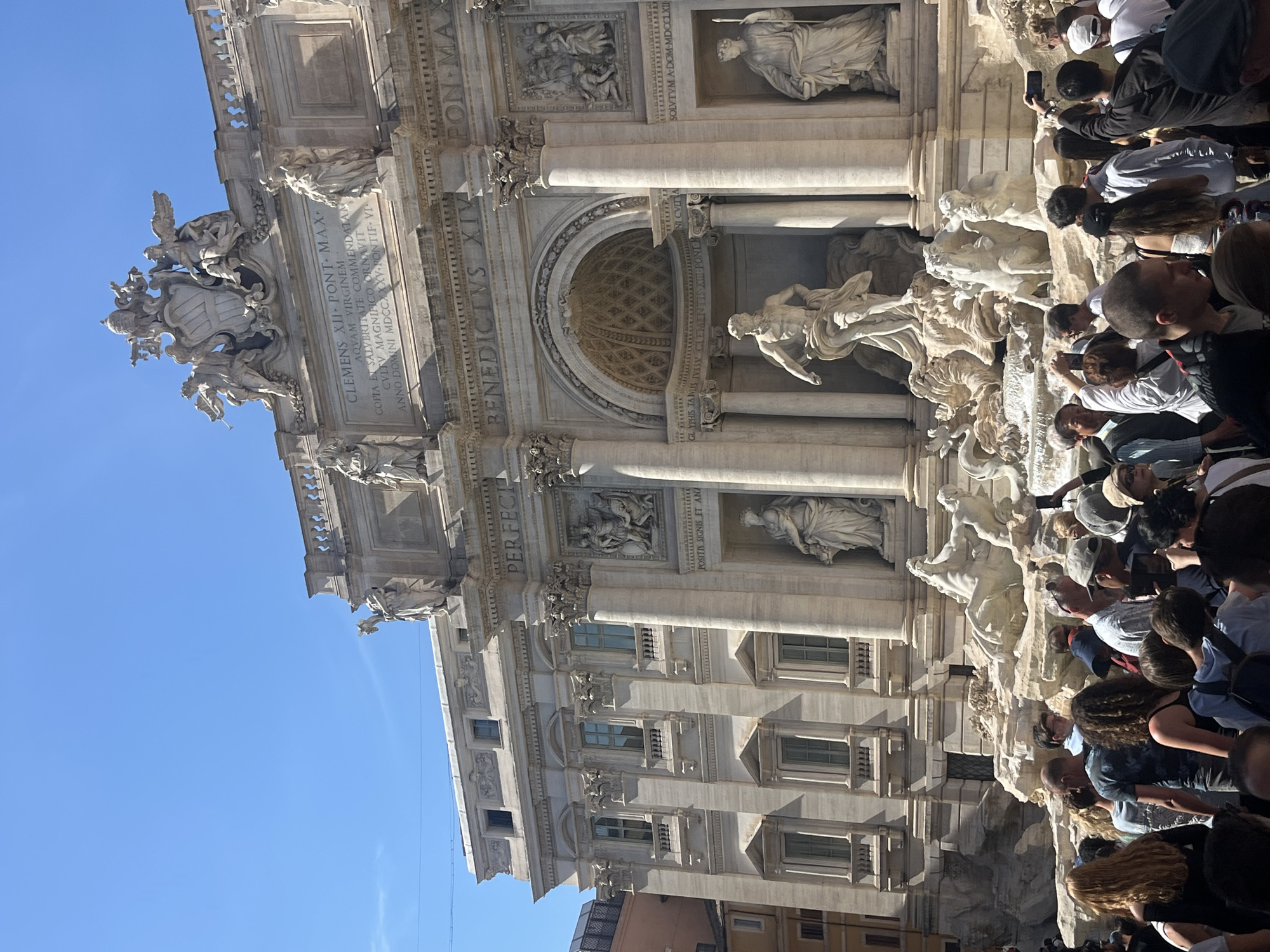 This image shows the Trevi Fountain in Rome, Italy, partially obscured by a large crowd of tourists. The fountain’s architecture is visible, including its statues, columns, inscriptions, and arched niches. The blue sky can be seen in the upper left corner. Many individuals are present, taking photos and interacting with the fountain. Buildings are visible in the lower right corner.