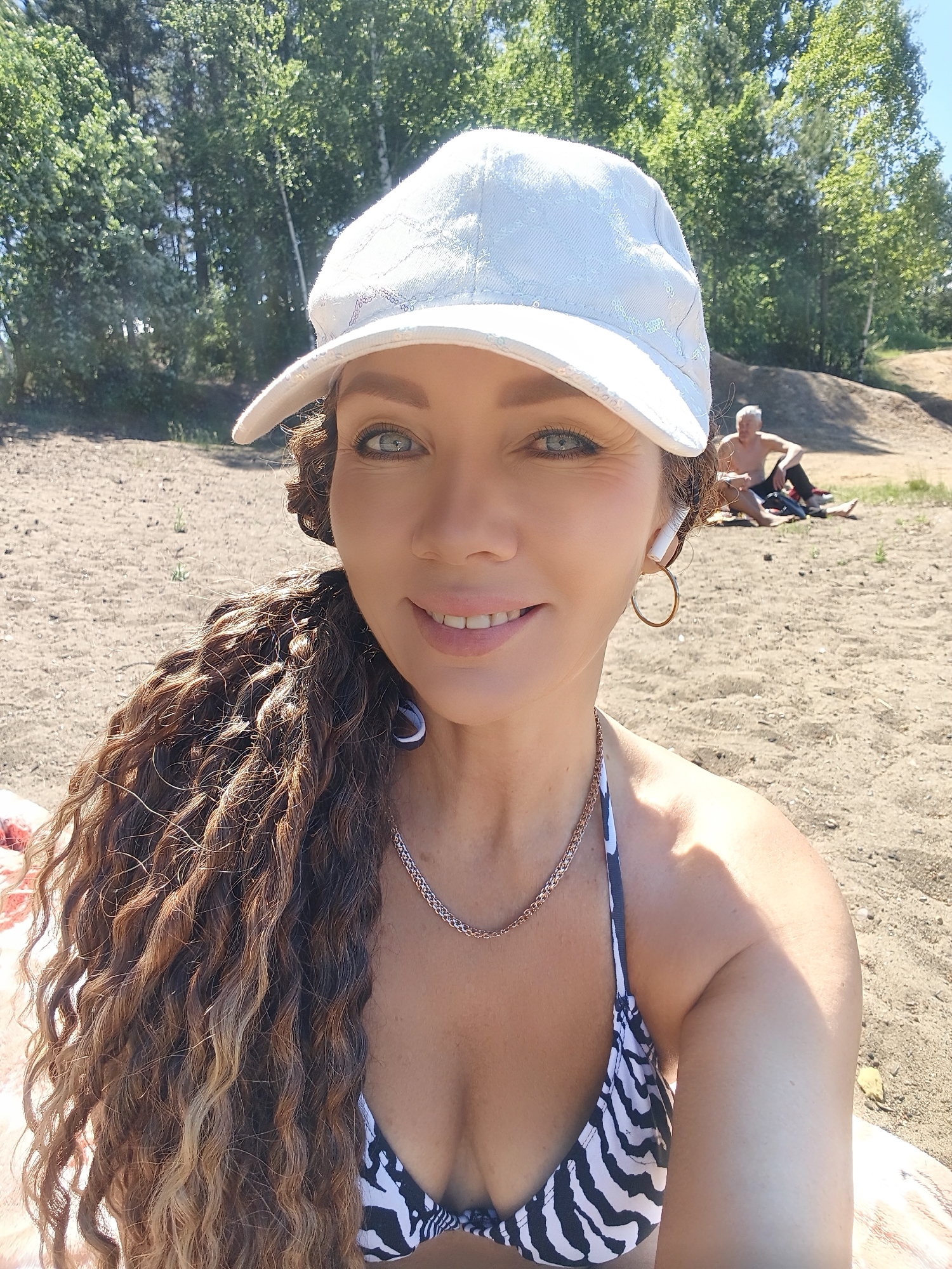 A woman in a white cap and a zebra-print bikini top is taking a selfie on a sandy beach. She has long, curly brown hair and is wearing hoop earrings and a necklace. In the background, a man is sitting on the sand among trees.