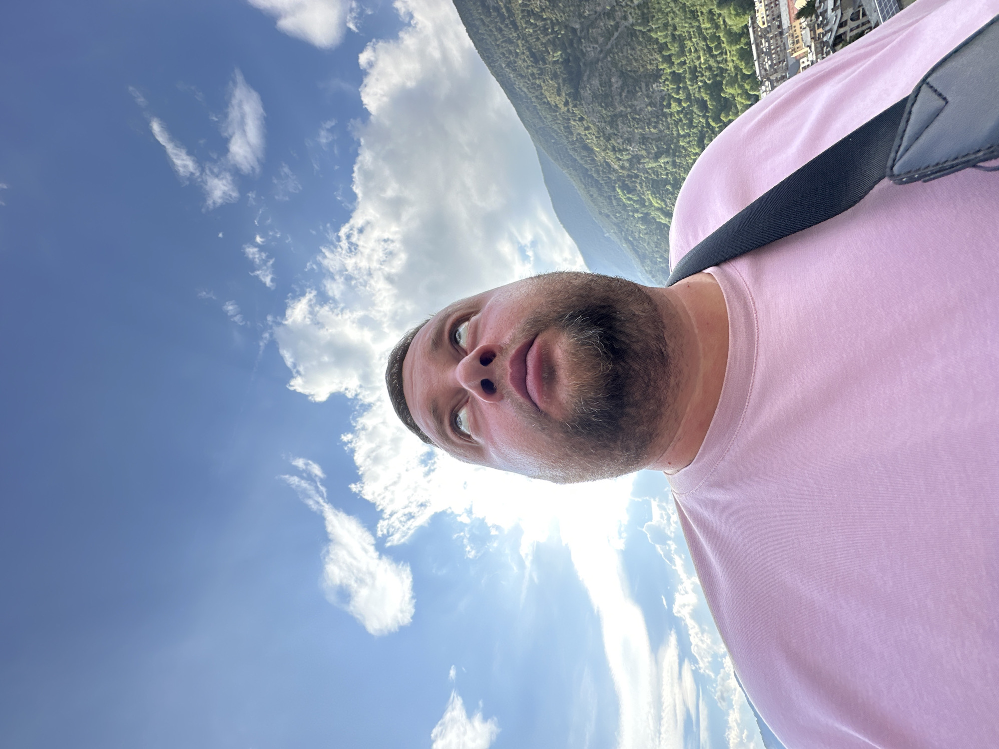 The image features a man's head and upper torso, positioned in the lower-central and right portion of the frame. He has short dark hair and a beard, wearing a light pink t-shirt with a black strap visible over his shoulder. His face is turned upwards and slightly to his left, with an observant or contemplative expression.
The background is dominated by a clear blue sky with scattered white, fluffy clouds, particularly prominent in the upper-left and central areas. A bright sun, emitting a strong lens flare, is visible in the upper-central part of the sky. To the right of the man's head, a large, steep mountain covered in dense green foliage extends from the lower-right upwards. Further up the mountain and towards the upper-right corner, several buildings of a town or village can be seen nestled in the landscape. The scene depicts a sunny day outdoors in a mountainous environment, consistent with the provided location context of Encamp, Andorra. There is no visible text in the image.