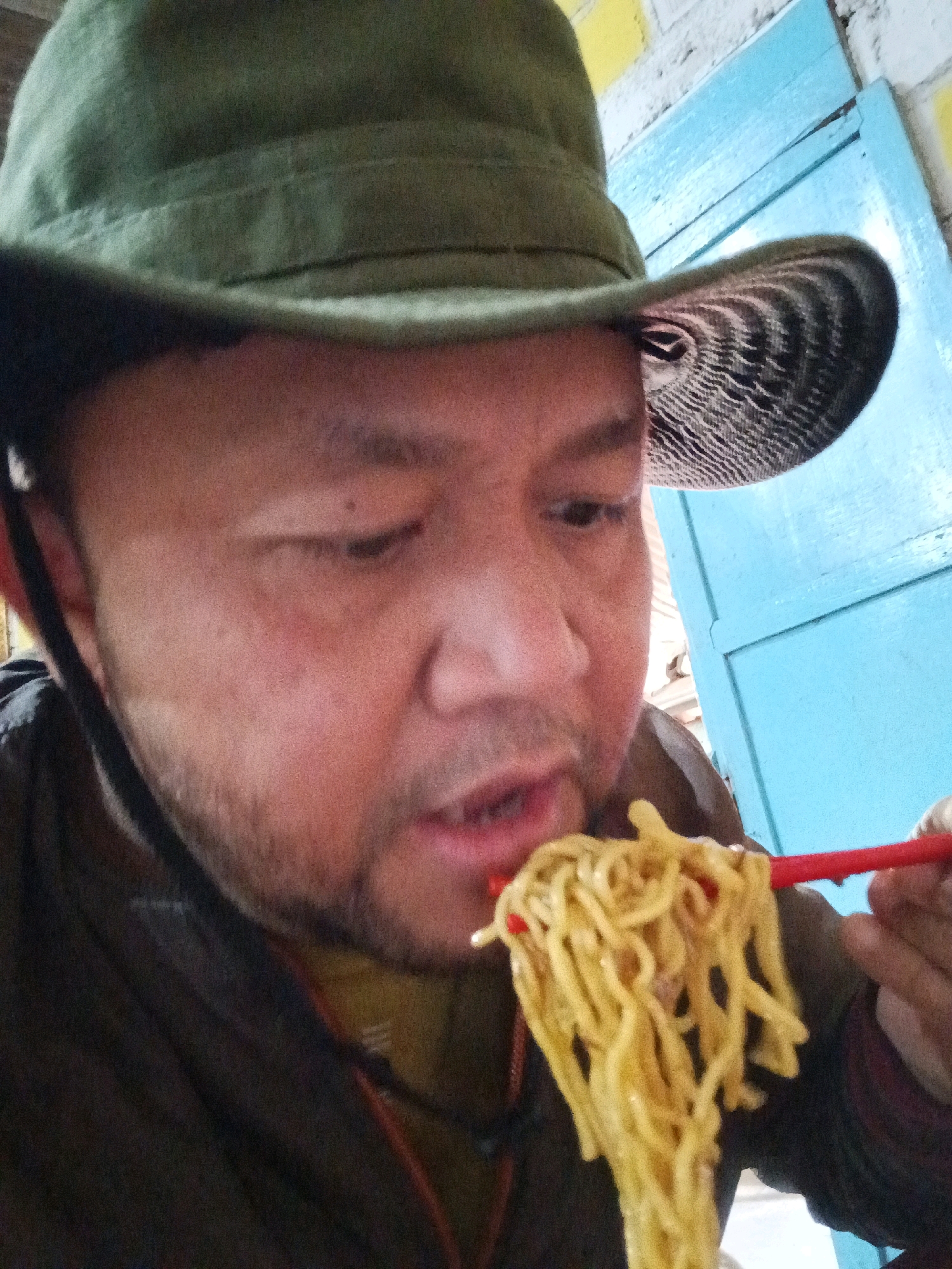 A close-up, low-angle selfie shows a person wearing a green bucket hat and a dark jacket eating noodles with red chopsticks. The person's face is partially visible, with their mouth open as they bring the noodles closer. The background features a blue wooden door and a brick wall with yellow and white paint. The scene is likely outdoors or in an open-air setting within Sraten, Indonesia, given the informal atmosphere and the type of food being consumed.
