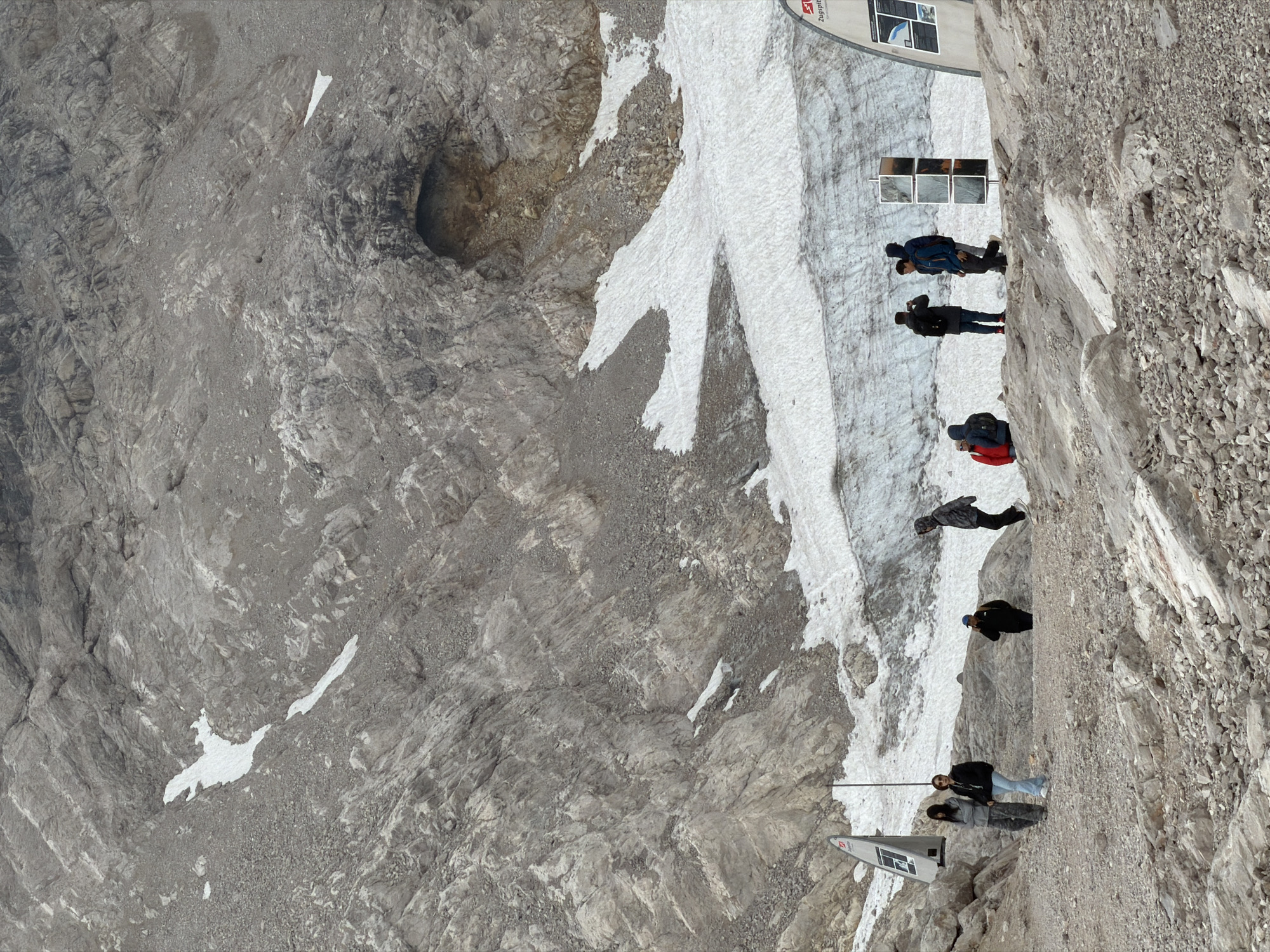 This image depicts a mountain landscape with sparse snow patches, rocky terrain, and a glacial area. Several individuals are visible, appearing to be tourists. Some are standing, others are walking, and one is taking a photo. They are dressed in casual clothing suitable for cool weather, with some wearing jackets and hoodies. The environment suggests a high-altitude location. The presence of informational signs and a structured viewing platform implies this is a designated tourist area. Based on the signs visible, specifically "Zugspitze," this scene is located at the Zugspitze, Germany's highest peak, which is near Garmisch-Partenkirchen.