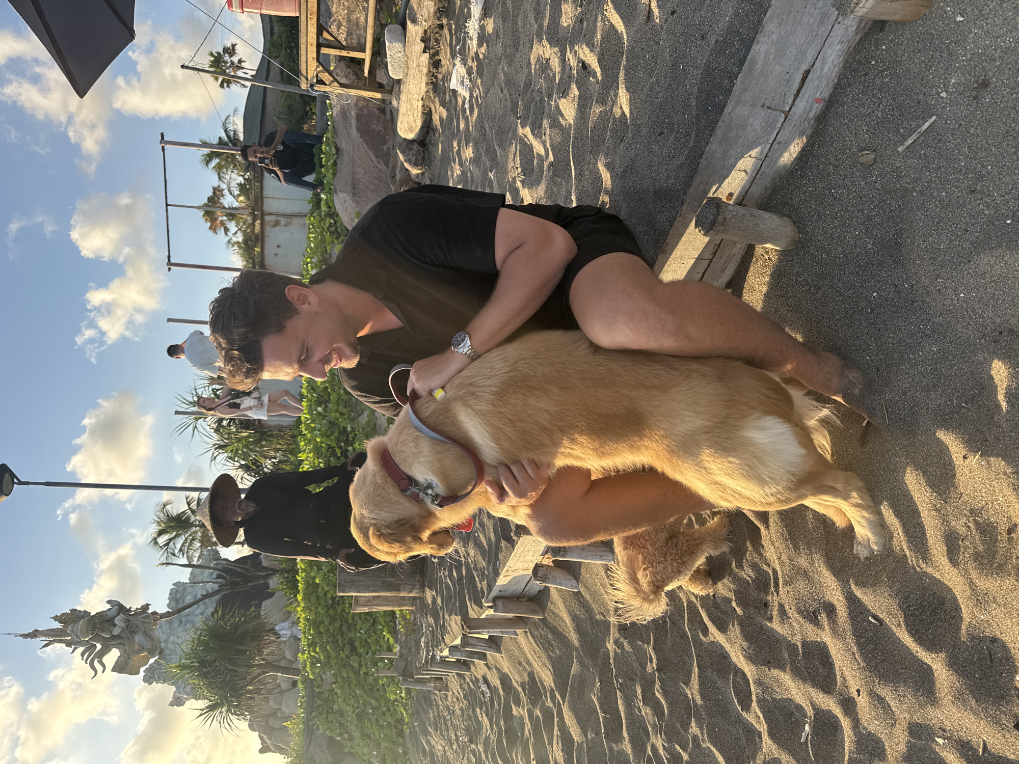 The photograph depicts a light-skinned man wearing a black t-shirt and dark shorts, seated on a wooden structure near the beach in an unknown location. The man is embracing a light-colored dog with a red and blue collar. The beach has dark sand with visible patterns. In the background, there is a sculpture, a person wearing a wide-brimmed hat, and vegetation. The sky is visible with some clouds.