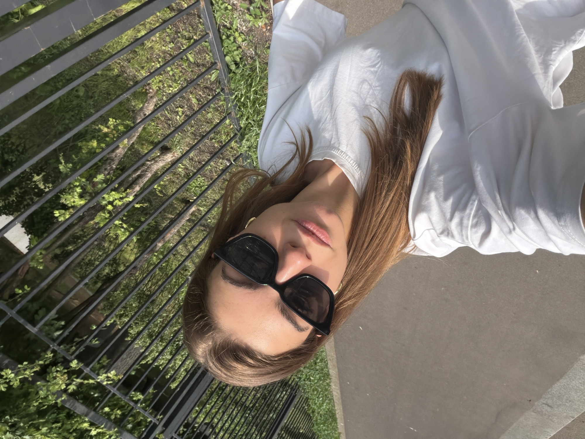 The image features a young woman outdoors in Moscow, Russia, captured in a rotated portrait under bright daylight. She is shown from the chest up, with long, light brown hair flowing over her shoulders and wearing a plain white t-shirt. Her face is partially obscured by black rectangular sunglasses, and her lips are slightly parted, conveying a neutral or serene expression. A gold hoop earring is visible on her left ear.
The woman is positioned next to a dark metal fence composed of horizontal bars, which appears diagonally across the frame due to the photo's orientation. Beyond the fence, a vibrant green landscape with grass, trees, and visible soil suggests a park or natural area. To her right, a light grey concrete surface, likely a sidewalk or pathway, is visible. The clear, strong sunlight indicates a sunny day, possibly midday or early afternoon. No specific activity is depicted beyond posing for the photograph, and no visible text is present.