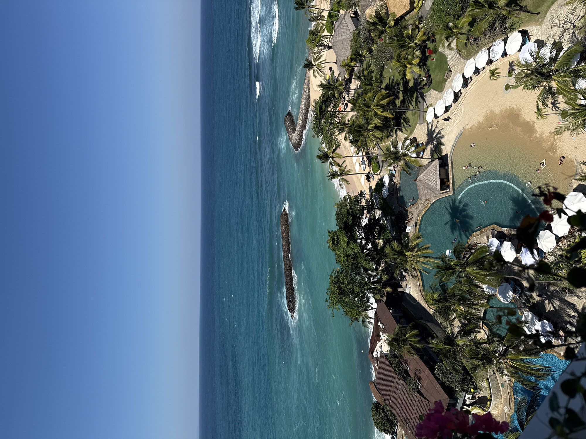 The image presents an aerial, high-angle view of a tropical resort situated along the coast in Kuta Selatan, Indonesia, under a clear, bright blue sky. Approximately the top quarter of the frame is occupied by the cloudless, light blue sky, which meets the horizon of the ocean. The ocean's color transitions from a deep blue at the horizon to a vibrant turquoise closer to the shore, exhibiting visible white-capped waves, particularly around two prominent, dark-colored rock breakwaters that extend in a curved fashion into the water from the coastline.
Below the ocean and sandy beach, the resort complex is characterized by abundant, lush green vegetation, dominated by numerous tall palm trees. The resort features multiple swimming pools; a large, irregularly shaped pool in the midground has a light sandy bottom, creating a shallow entry zone that mimics a beach. Approximately ten individuals are visible within this pool, swimming, wading, or floating on inflatables. Another segment of a pool is partially visible in the lower-left corner.
Scattered throughout the resort grounds are several low-rise buildings with dark brown, tiled roofs, blending with the natural environment. A small, round, thatched-roof structure is positioned adjacent to the large pool. White lounge chairs and white beach umbrellas are systematically arranged on the sandy areas around the pools and near a section of natural beach. A paved pathway runs alongside a row of these umbrellas, where one individual is observed walking. In the immediate lower-left foreground, blurred pink flowers and green foliage indicate the viewpoint is from an elevated balcony or terrace. The scene is illuminated by direct sunlight, casting distinct shadows and suggesting a midday to early afternoon timeframe.