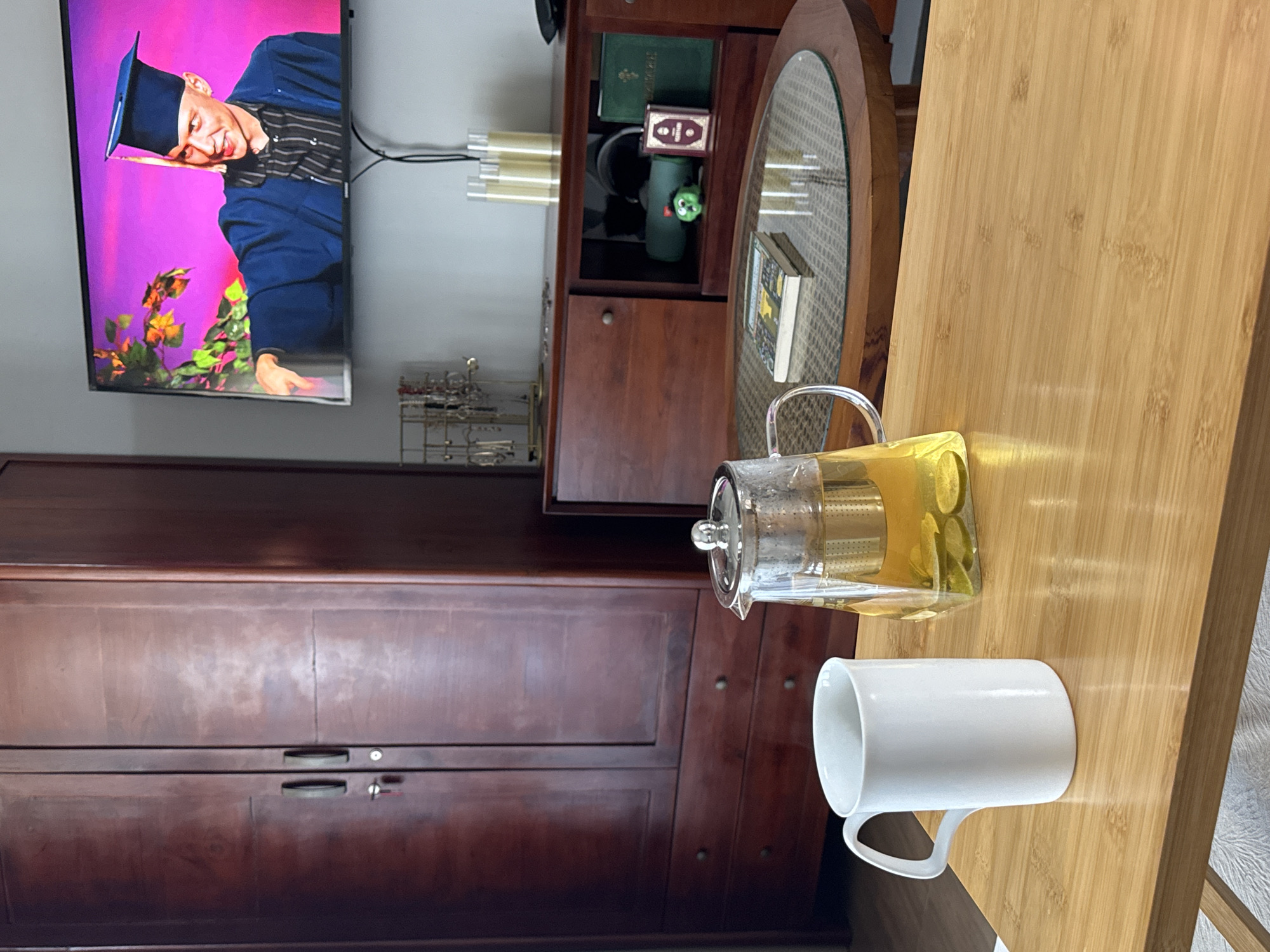 In the foreground, a white ceramic mug sits on a wooden coffee table. Next to the mug is a glass teapot filled with yellow liquid and slices of lemon. In the background, a large wooden wardrobe stands to the left, and a television screen displays a person in a graduation cap and gown against a vibrant pink background. A wooden cabinet with shelves is visible below the television. The scene appears to be an indoor living space. There is no indication that this scene is located in North Kuta, Indonesia.