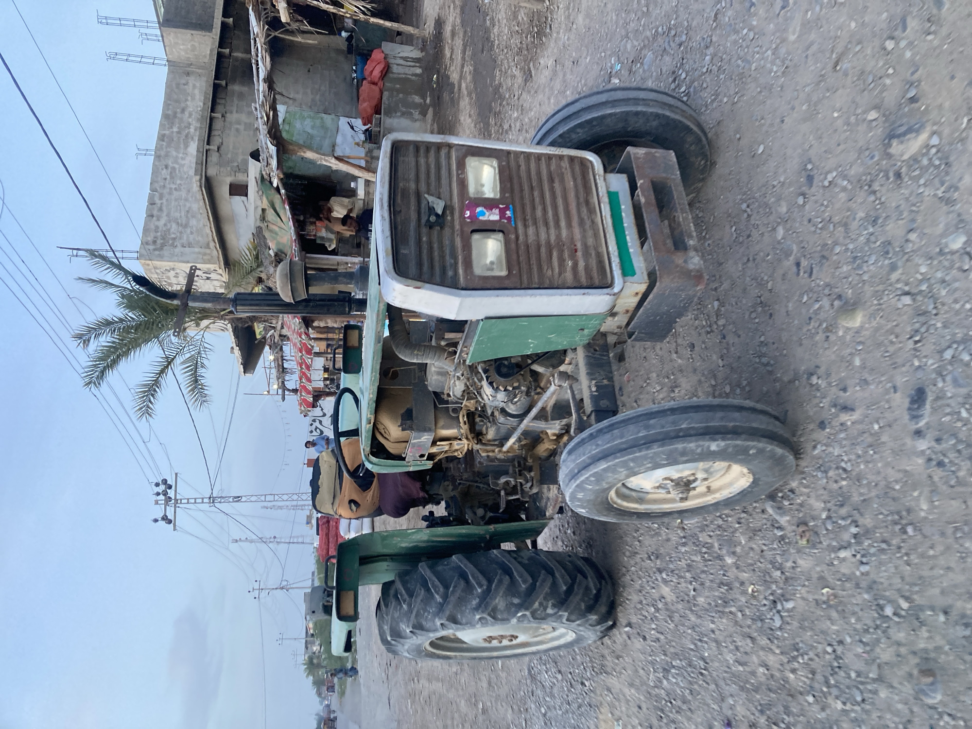 The image depicts a green and white tractor parked on a gravel road in Uthal, Pakistan. The tractor is positioned perpendicular to the road, angled slightly towards the right. The surrounding environment includes buildings of varying construction styles, some appearing to be in a state of disrepair. A palm tree is visible in the background, along with electrical power lines and poles. Other vehicles and pedestrians are present in the distance along the road.