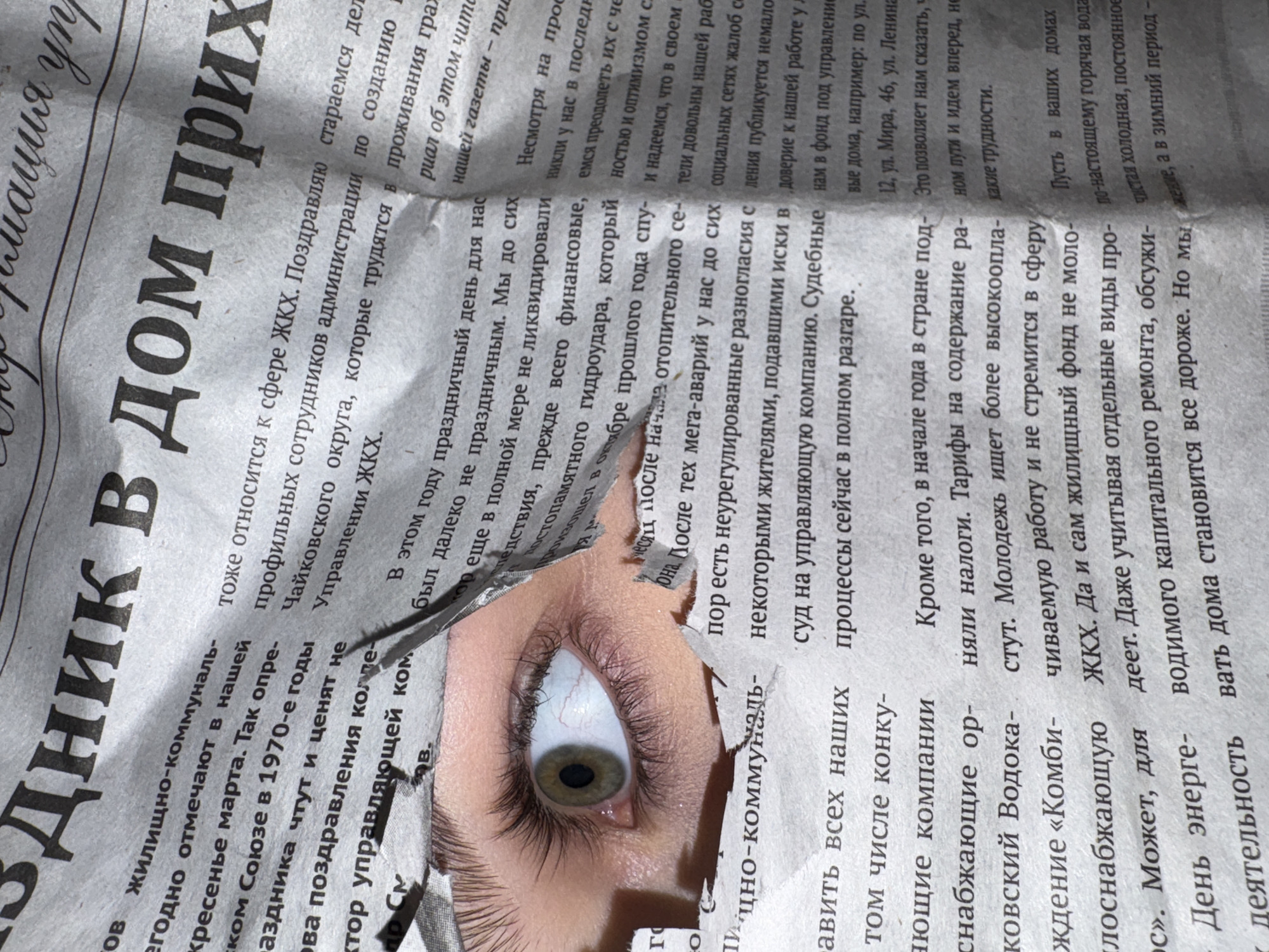 The image shows a close-up of a person's eye peering through a torn newspaper. The eye has a green iris. The surrounding skin is pale and framed by dark eyelashes. The newspaper itself is printed in Cyrillic script, suggesting it is Russian. Text is visible around the tear. Parts of the newspaper mentions the city of Chaikovsky, Russia.