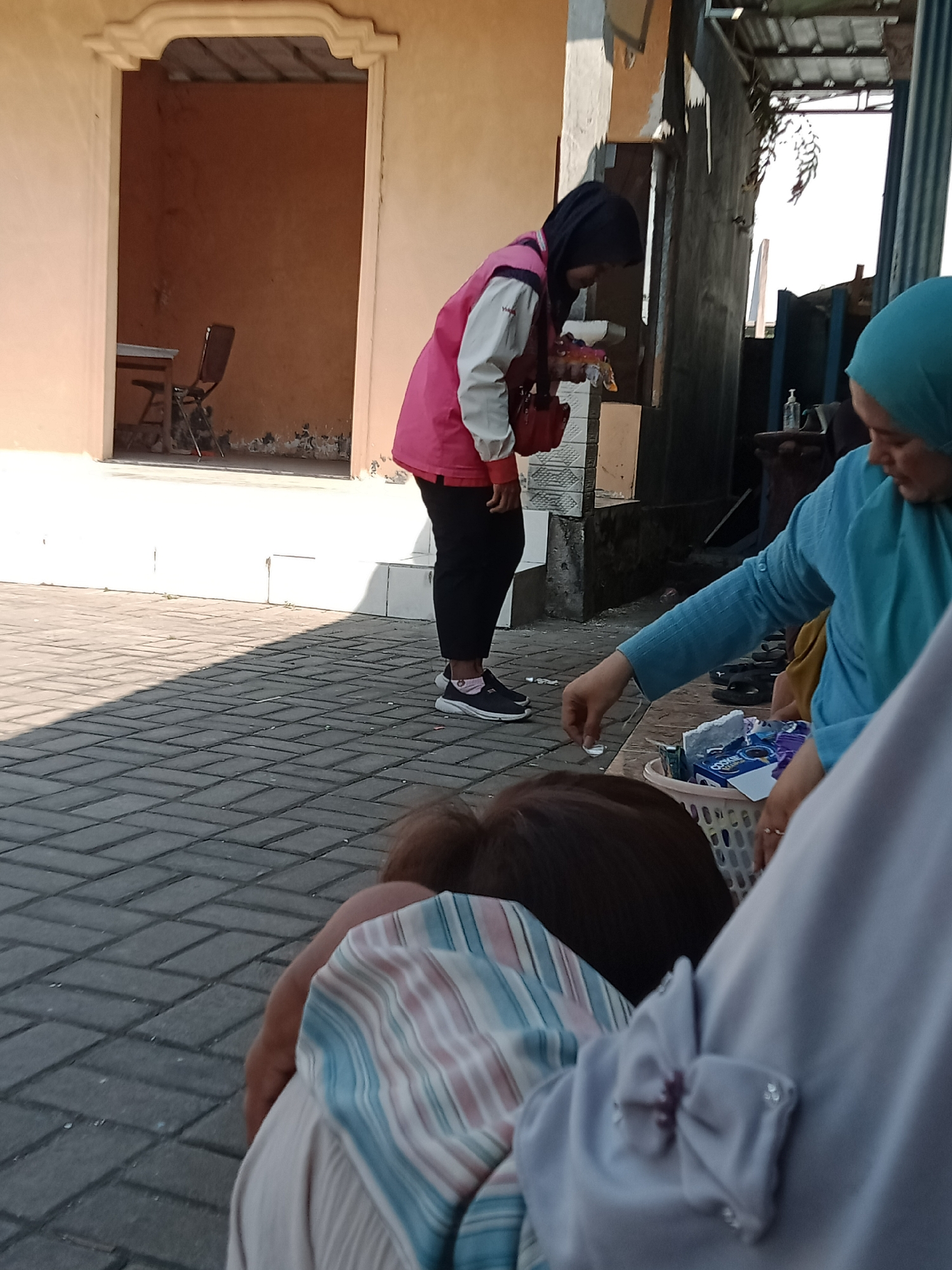 In Simogirang, Indonesia, a young woman wearing a pink and white jacket, a black headscarf, and black pants stands near a doorway. She is leaning forward, holding a small object in her right hand, and looking down. In front of her, a woman in a blue headscarf and blue top is reaching into a white basket containing packaged snacks. Another person, partially visible in the foreground, is wearing a light purple garment with a decorative bow. The ground is paved with grey interlocking bricks, and a shadow from an unseen structure falls across it. Behind the young woman, a doorway reveals an interior with an orange wall, a table, and a chair.