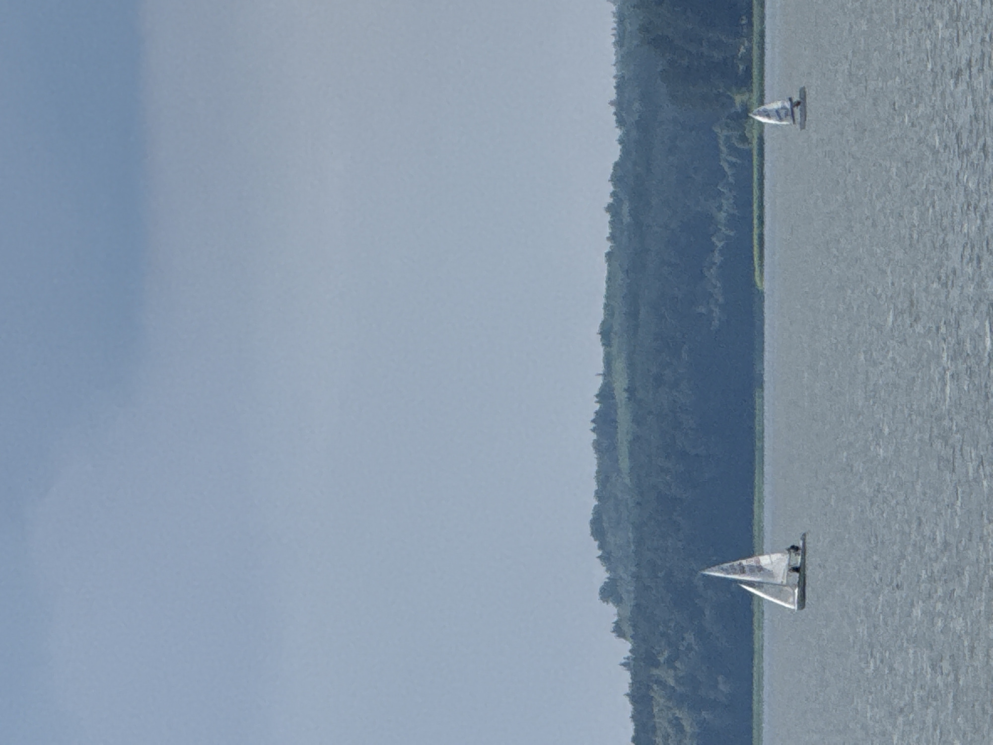 The image depicts a serene scene on a body of water, likely a lake or a wide river, with two sailboats navigating the calm surface. The sky above is a muted blue, with overcast conditions contributing to a soft, diffused light. In the background, a lush, tree-covered hill forms a natural backdrop, its dense foliage a deep green.
The foreground is dominated by the water, its surface textured by gentle ripples. To the left, a white sailboat with a triangular sail is prominently featured. Two figures are visible on board, appearing to be engaged in sailing. Further to the right, a smaller sailboat, also with a white sail, is visible in the distance. The horizon is defined by the meeting of the water and the distant treeline.
The location is identified as Votkinsk, Russia, a city known for its picturesque natural surroundings and its connection to the composer Pyotr Ilyich Tchaikovsky. This particular scene evokes a sense of tranquility and outdoor recreation, common to lakeside or riverside settings. The absence of urban structures suggests a natural or park-like area within or near the city. The overall impression is one of peaceful exploration and enjoyment of the aquatic environment.