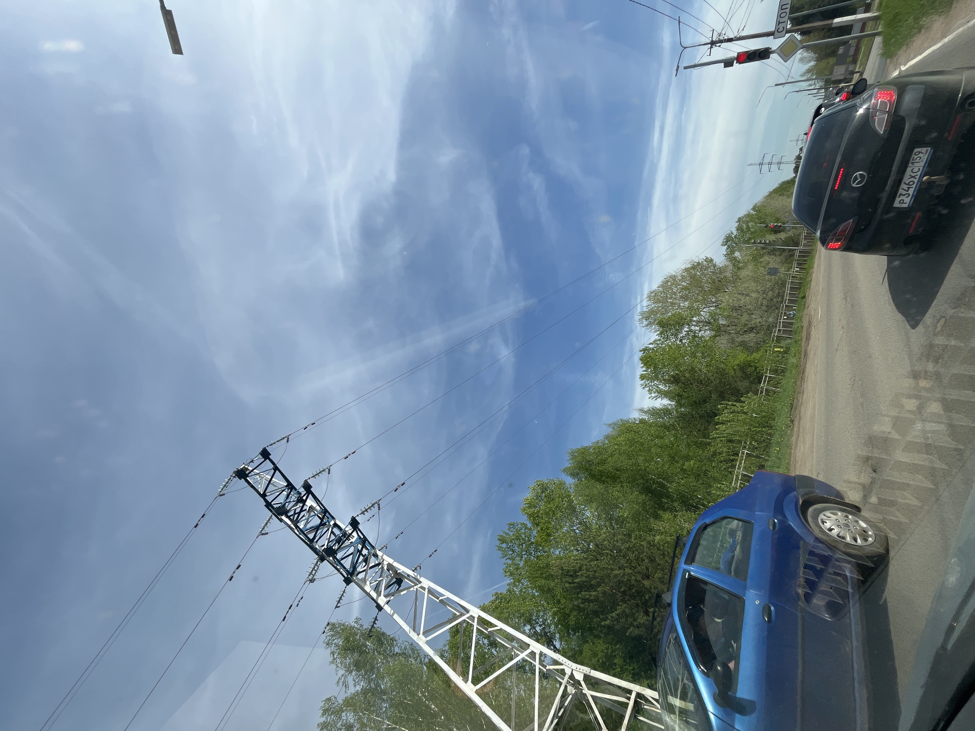 The image depicts a street scene in Chaikovsky, Russia. A blue car and a black car are on the road. A traffic light is visible with a red light illuminated. Power lines and a white and blue power line support structure are prominent in the foreground. Trees and vegetation line the roadside. The sky is blue with scattered clouds. The black car has the license plate P346XC 159. The image appears to be taken from inside a vehicle.
