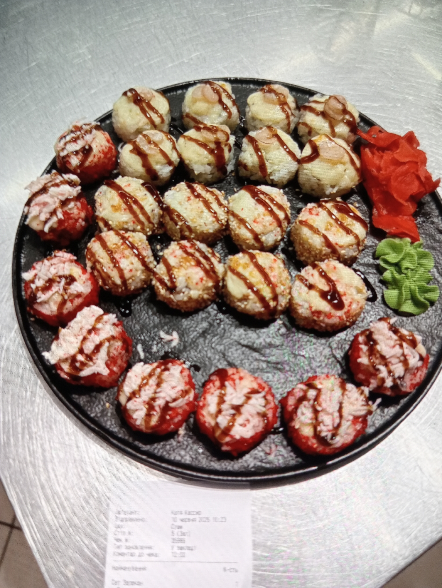 A circular black plate containing an assortment of sushi and sashimi is presented on a metallic surface. The sushi pieces are arranged in concentric circles. The outer layer consists of red-colored sushi, topped with a white filling and drizzled with a dark sauce. The inner layers feature lighter-colored sushi, some with a creamy white topping and a sprinkling of sesame seeds, also drizzled with a dark sauce. A small pile of bright pink pickled ginger and a mound of green wasabi are situated to the right of the sushi. The background is a textured metallic surface, possibly a countertop. A receipt is visible at the bottom left of the image, partially obscuring the metallic surface. The receipt contains Cyrillic text and numerical data. The overall lighting suggests an indoor setting.