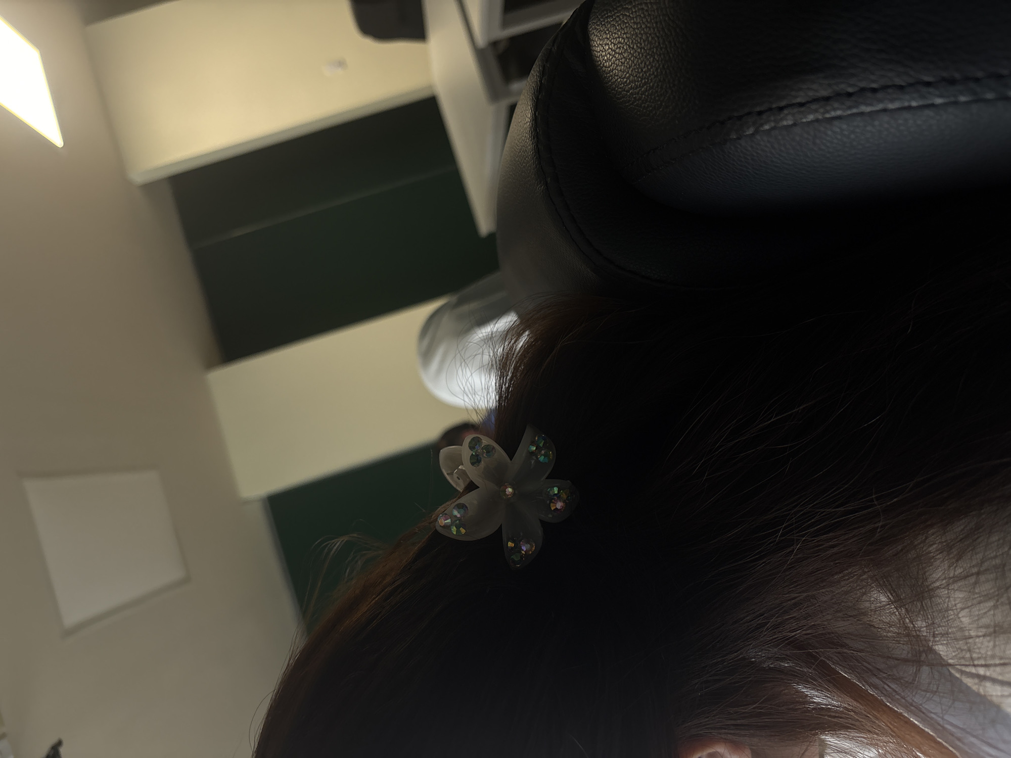 The image shows the back of a person's head with dark brown hair. A light-colored, flower-shaped hair clip is visible in the hair. The person appears to be seated in a chair with a black leather headrest. The background includes a wall with a two-toned pattern, and a light source that appears to be a fluorescent light fixture. It is not possible to determine the location in the city of Puebla, Mexico.