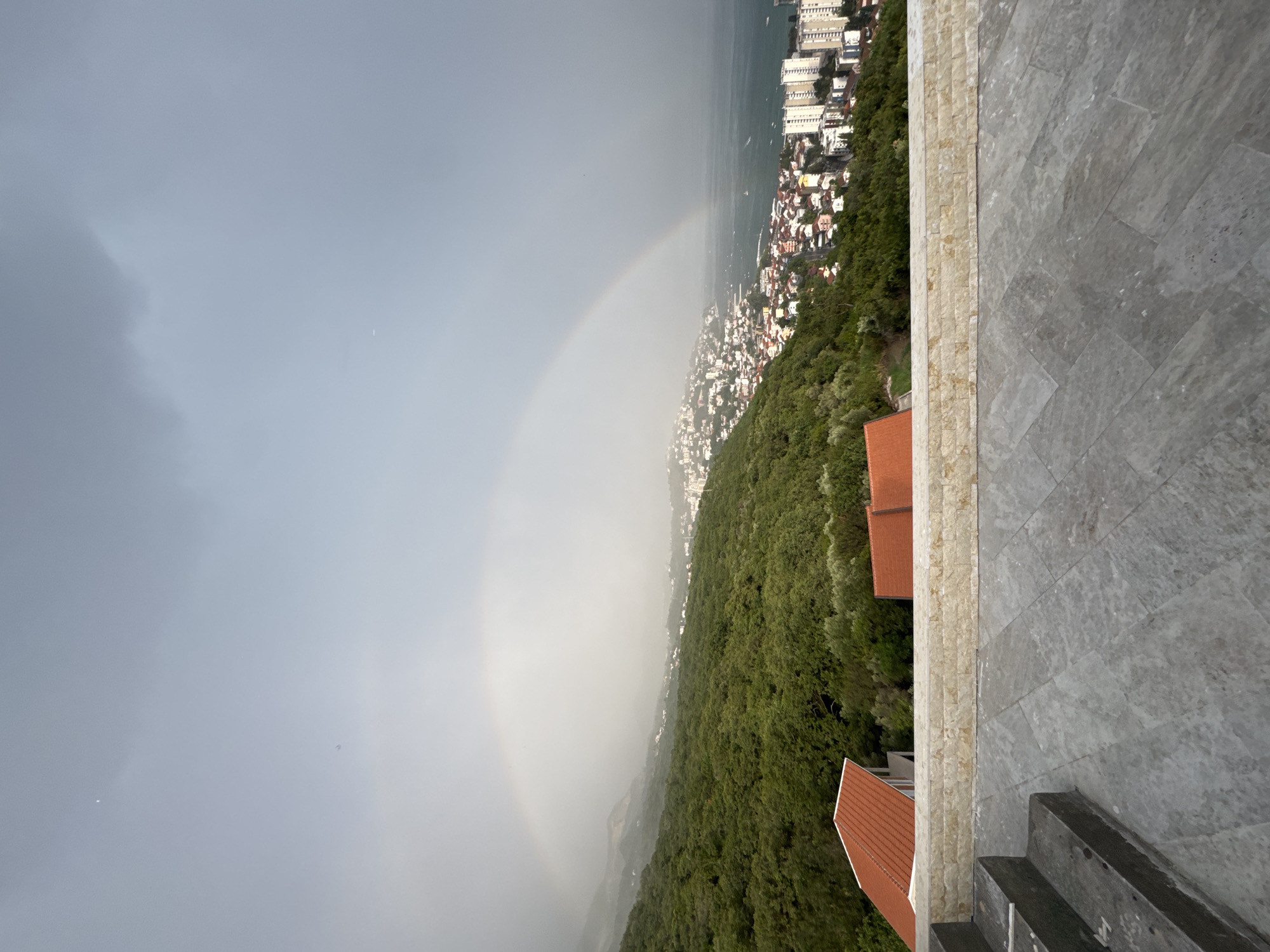 The image depicts a panoramic view from an elevated vantage point, showcasing a coastal city nestled amongst lush green hills and bordering a calm body of water. A faint rainbow arcs across a hazy, overcast sky, suggesting recent or ongoing precipitation. The city is characterized by a dense cluster of buildings with white facades and terracotta roofs, rising up the hillsides and extending towards the waterfront. Several larger, modern buildings are visible along the coast.
In the foreground, a modern stone patio area with steps leads down towards the cityscape. The patio is paved with large, grey stone tiles and a border of smaller, lighter-colored rectangular stones. The overall scene suggests a temperate climate and a scenic, possibly Mediterranean, coastal location. The time of day appears to be late afternoon or early evening, given the soft, diffused light and the overcast conditions. There are no people or specific objects of focus in the image, other than the natural and architectural elements of the landscape. The mood conveyed is one of serene beauty, with the rainbow adding a touch of wonder to the scene. No visible text is present in the image. The location context provided is Sušćepan, Montenegro.