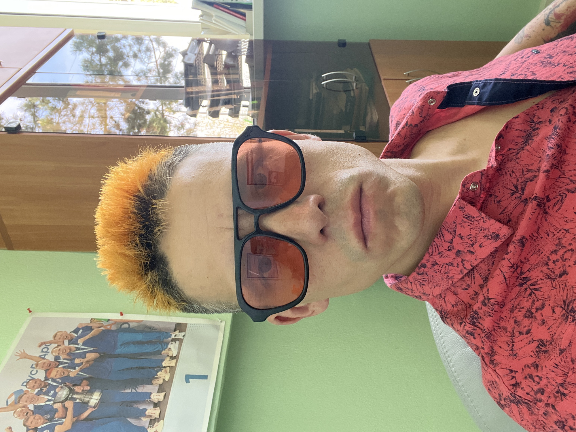 A man with orange hair is wearing orange-tinted sunglasses and a pink patterned shirt. He is in a room with a light green wall and a calendar featuring a sports team holding a trophy. The calendar has the number "1" on it and the text "РУСАЛ Арт" (RUSAL Art). The room appears to be an office setting, as there are cabinets and a window visible in the background. While the text on the calendar refers to RUSAL, a Russian aluminum company, I cannot confirm the location with certainty as being in Chaikovsky, Russia, from this image alone.