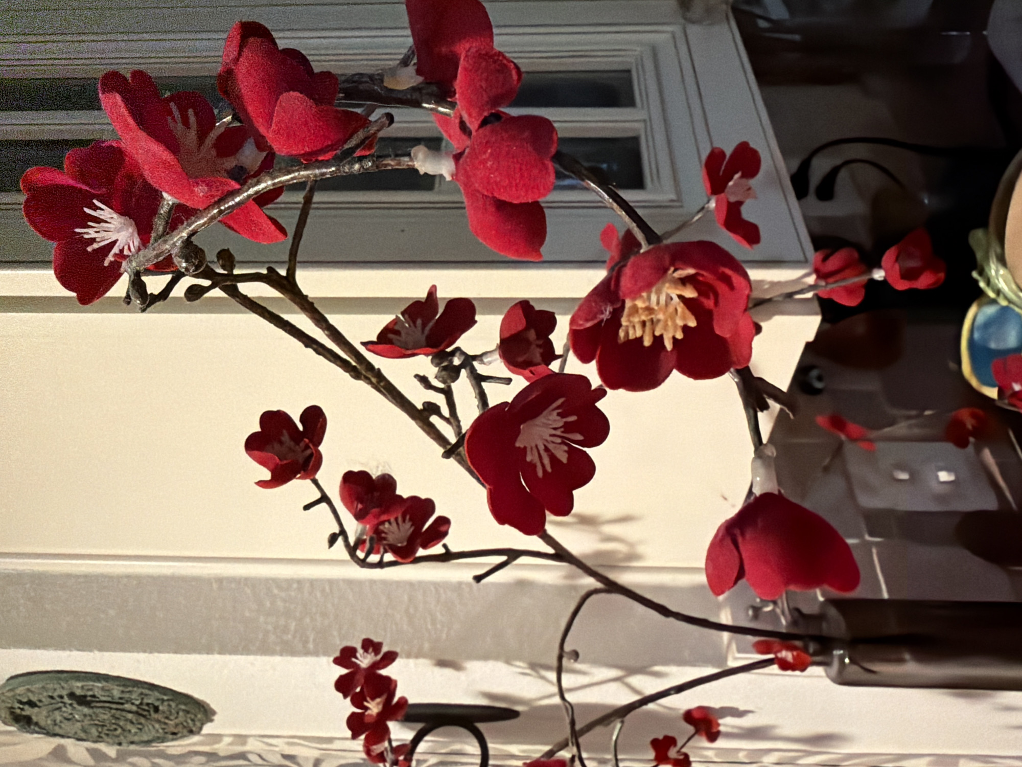 This is a close-up, eye-level, indoor shot of a decorative artificial flower arrangement. The flowers are a vibrant red with white centers and are arranged on thin, dark branches. Some of the flowers are in full bloom, while others are buds. The arrangement appears to be placed in a dark, opaque vase that is partially visible at the bottom left of the frame. In the background, a cream-colored wall is visible, with a decorative embossed object hanging on it to the left. A window with panes is seen on the right side of the background. The lighting suggests an indoor setting, possibly a home or a commercial space, with the light source casting shadows. The focus is sharp on the flowers in the foreground, with the background slightly blurred. There are no people or animals visible in the image.
