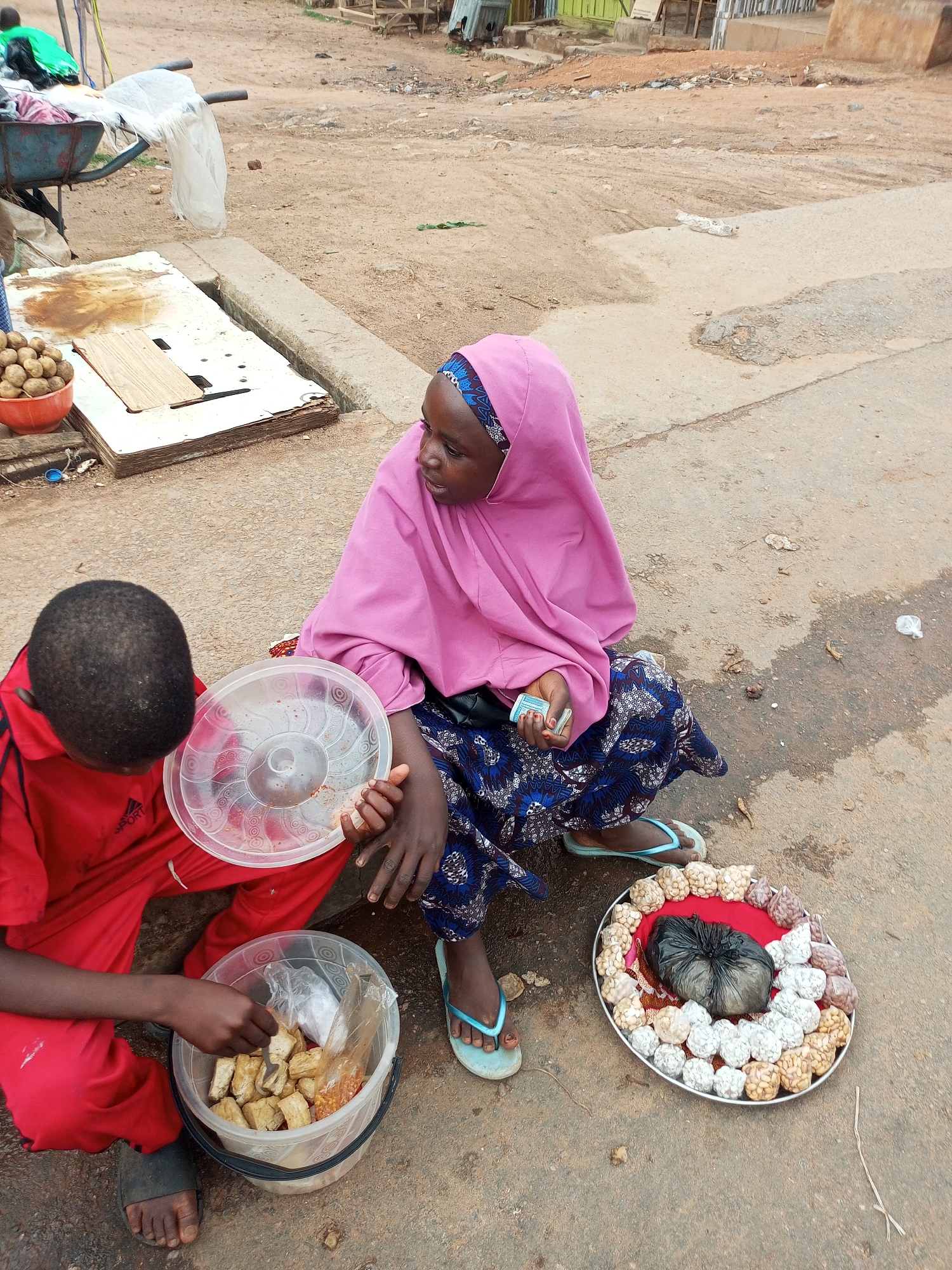 In Tempure, Nigeria, a young girl in a pink hijab and a patterned blue skirt sits on the ground, her bare feet wearing light blue flip-flops. She holds a small stack of bills in her right hand. Beside her, a young boy in a red tracksuit is crouched down, reaching into a clear plastic bucket filled with what appear to be pieces of fried food, possibly tofu or plantain, and some smaller items. A clear plastic lid rests on the ground near him. To the girl's right, a silver tray is arranged with small, round food items, possibly nuts or candies, in various colors and textures. A black plastic bag is placed in the center of the tray. The ground is dusty and uneven, with patches of concrete and dirt. In the background, a wheelbarrow is partially visible, laden with fabrics and a white plastic bag. The scene suggests a street-side market or a vendor selling food.