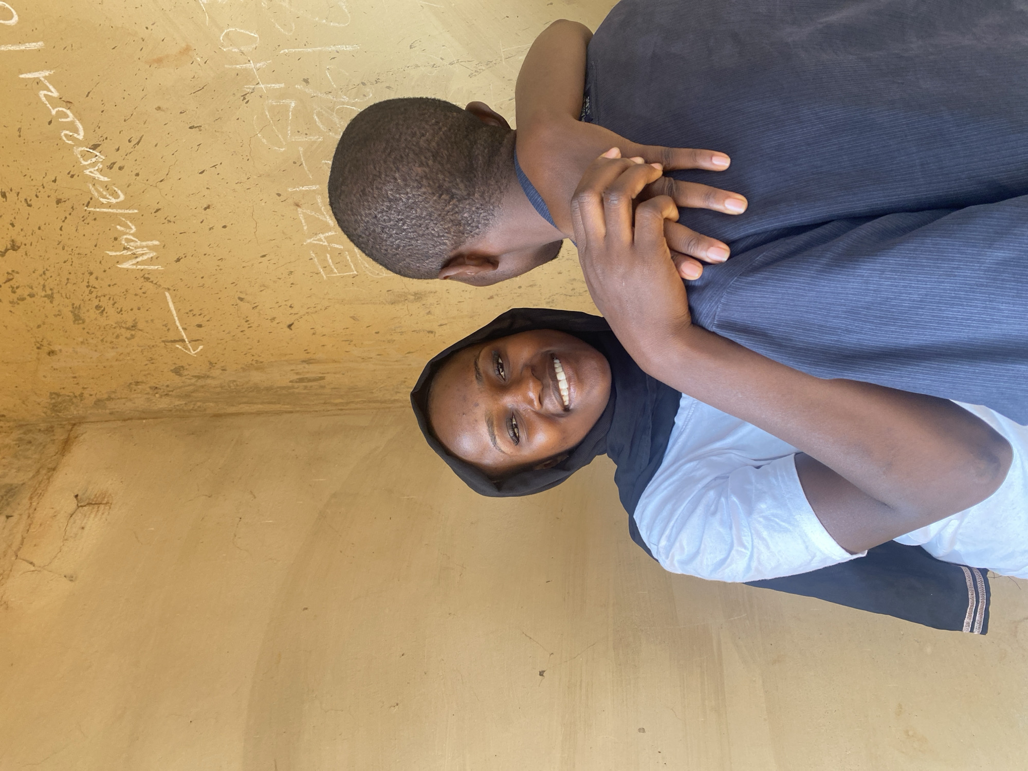 The image captures two individuals, a woman and a man, posing in an embrace against a plain, light brown wall. The woman, on the right, faces the camera with a wide, joyful smile. She is wearing a black head covering (hijab) and a white short-sleeved top. Her hands are clasped over the chest area of the man, who is positioned to her left, largely with his back to the camera. The man has short, dark hair and is wearing a dark, possibly navy or black, collared shirt. His arm appears to be around her, suggesting a close and affectionate relationship.
The setting appears to be an indoor or covered space, characterized by the simple, unadorned wall. The wall shows signs of age and wear, with minor cracks, dust, and scattered specks. White chalk writing is visible on the upper part of the wall. The most prominent text reads "NPC/EAD 2021" with an arrow pointing downwards, and another inscription below it appears to be "EAZI" or "EAZI-1". These markings could be related to census or demographic activities, such as those conducted by the National Population Commission (NPC) for Enumeration Area Demarcation (EAD) in areas like Kuru Dungus, Nigeria. The lighting is soft and even, indicating either diffused natural light or an indoor setting without direct harsh sunlight. The overall impression is one of happiness and connection in a humble, functional environment.