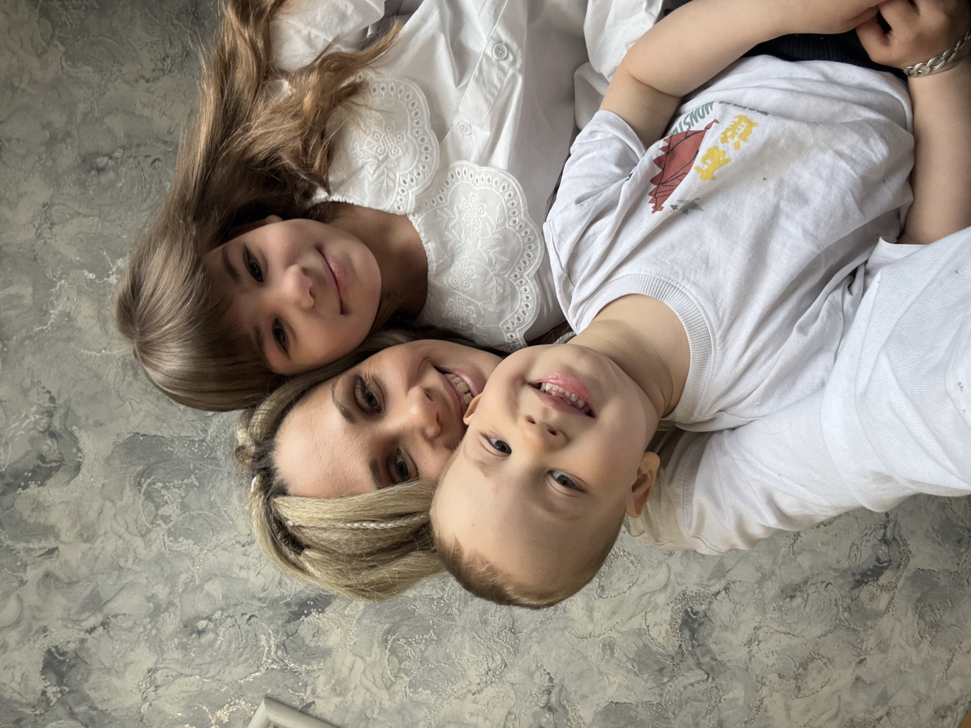 A top-down, medium shot shows a woman, a girl, and a boy lying on a gray and white patterned surface. The woman has blond hair, and is smiling. The girl has brown hair with bangs. The boy has short, light brown hair and is also smiling. They are all wearing light-colored clothing. The scene appears to be inside, possibly in Rusanovo, Russia, as specified in the prompt.