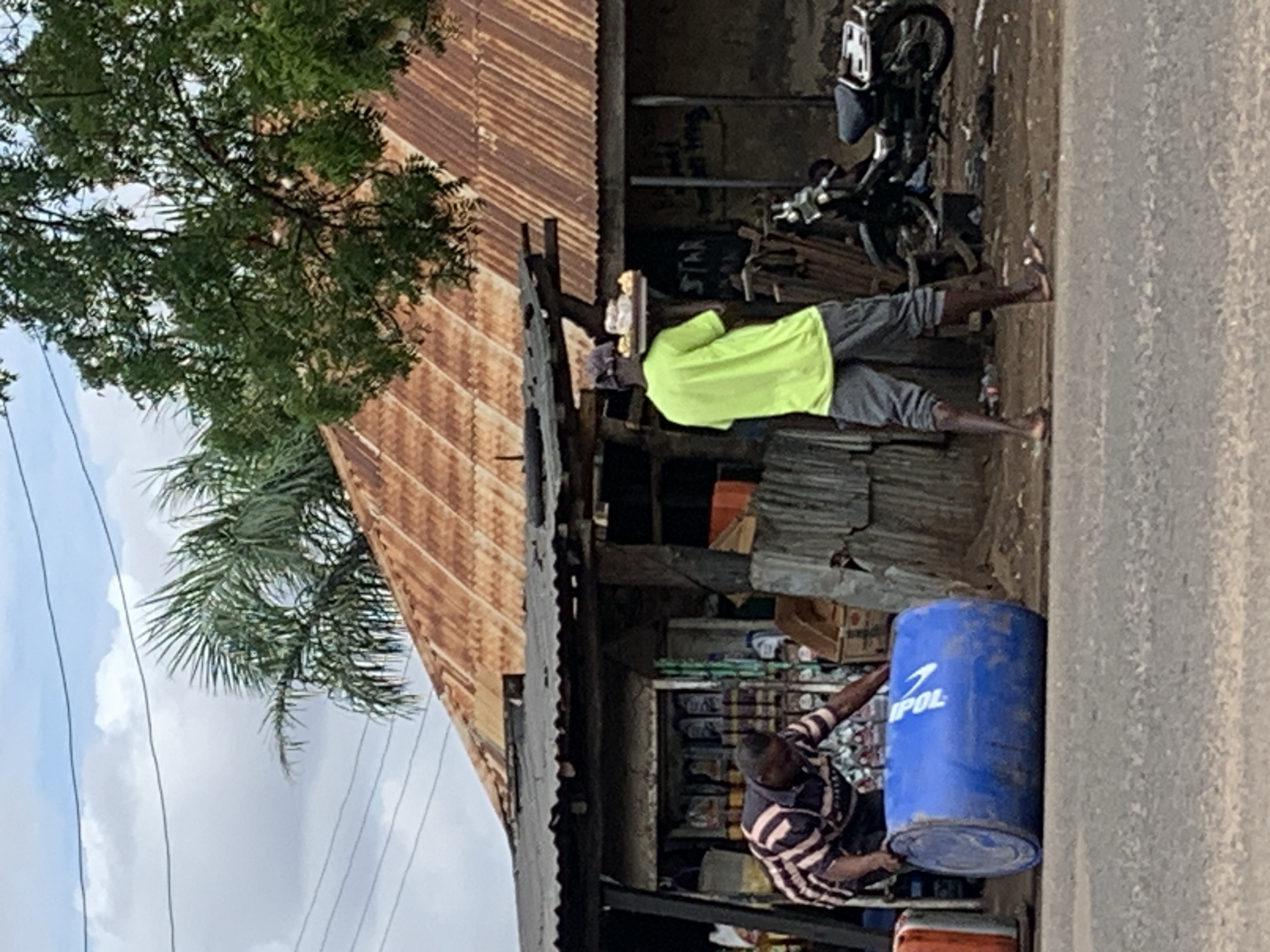 The image depicts an outdoor scene in Shendam, Nigeria, under a partly cloudy sky. In the foreground, a grey paved road stretches across the lower portion of the frame.
On the left side of the road, a male individual with dark skin, wearing a horizontal striped shirt (dark and light beige/pink stripes) and dark trousers, is bent over, pushing a large blue plastic barrel lying on its side. The barrel has white lettering, partially visible as "POL".
Towards the right, a second male individual with dark skin is walking away from the viewer. He wears a bright lime green short-sleeved t-shirt, grey capri-length shorts/trousers, and a dark baseball cap. He is balancing a flat, dark-colored tray on his head, laden with multiple light-colored items, consistent with baked goods or pastries. He wears open-toed footwear.
Behind the two individuals, on an unpaved dirt shoulder, stands a small, open-front structure resembling a shop or kiosk, featuring a dark, corrugated metal roof. Shelves stocked with various products are visible inside this structure. Further back and to the right, a dark-colored motorcycle is parked on the dirt.
The background features several buildings with steeply pitched, rusted corrugated iron roofs. Lush green foliage from various trees, including palm fronds on the left and dense leafy trees on the right, partially obscures these structures. Multiple dark power lines crisscross the light blue and white cloudy sky in the upper left quadrant of the image.