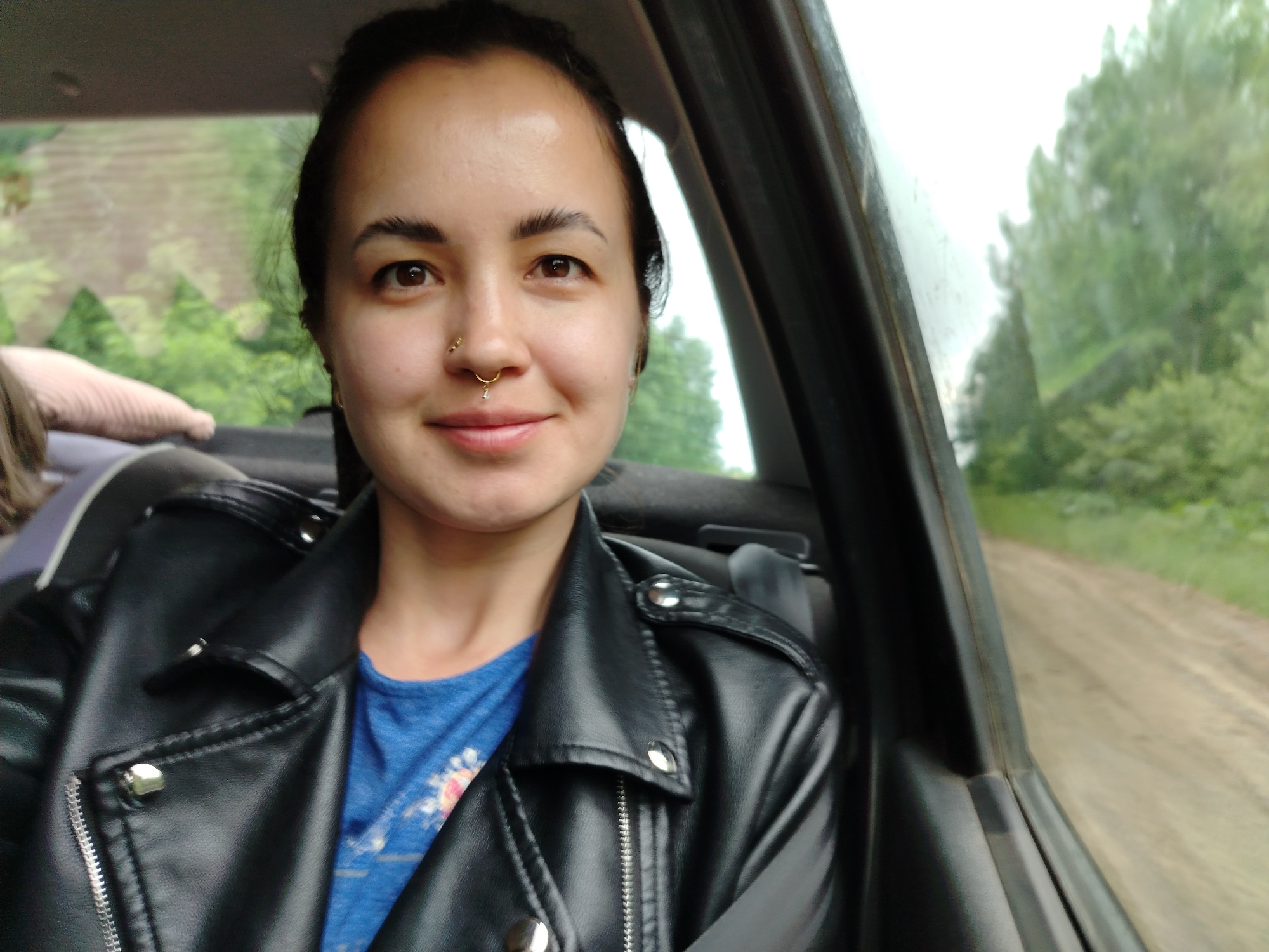The image appears to be a selfie taken inside a moving vehicle. The subject is a woman with brown eyes and dark hair pulled back from her face. She has a nose ring. She is wearing a blue t-shirt and a black leather jacket, and a seatbelt is visible across her chest. Outside the car window, there is a blurry view of green trees and a dirt road, suggesting a rural or wooded area. The photograph was taken somewhere near Krasnoyarsk, Russia.