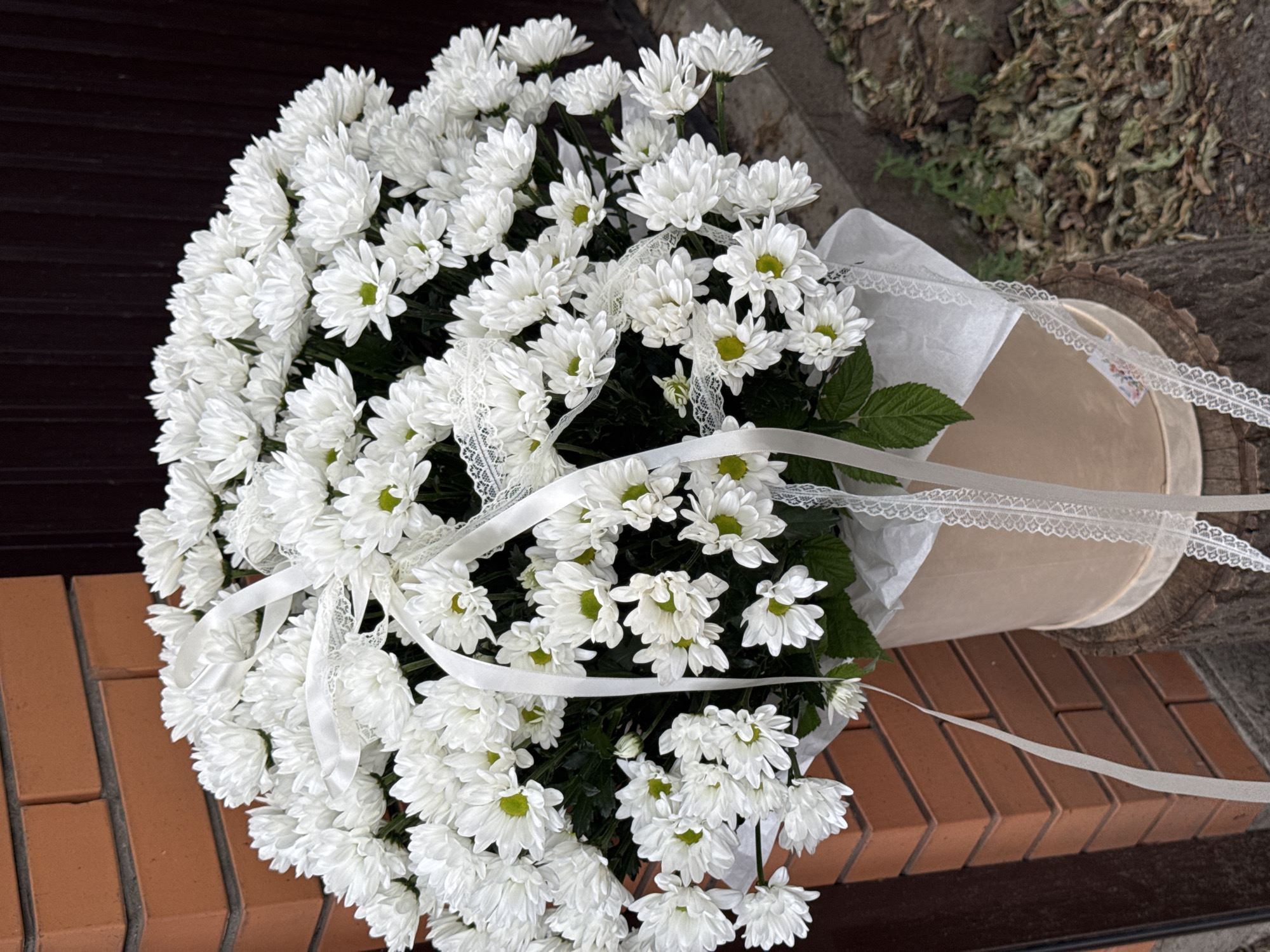 A large bouquet of white chrysanthemums with green centers is presented in a light beige hatbox, sitting on a tree stump. The bouquet is adorned with white satin ribbons and delicate white lace trim. The background features a brick wall on the left and a dark wooden fence or door on the right. The ground is covered with dry leaves and some green vegetation. The image is captured outdoors in what appears to be a residential or garden setting. There are no people or animals in the image.