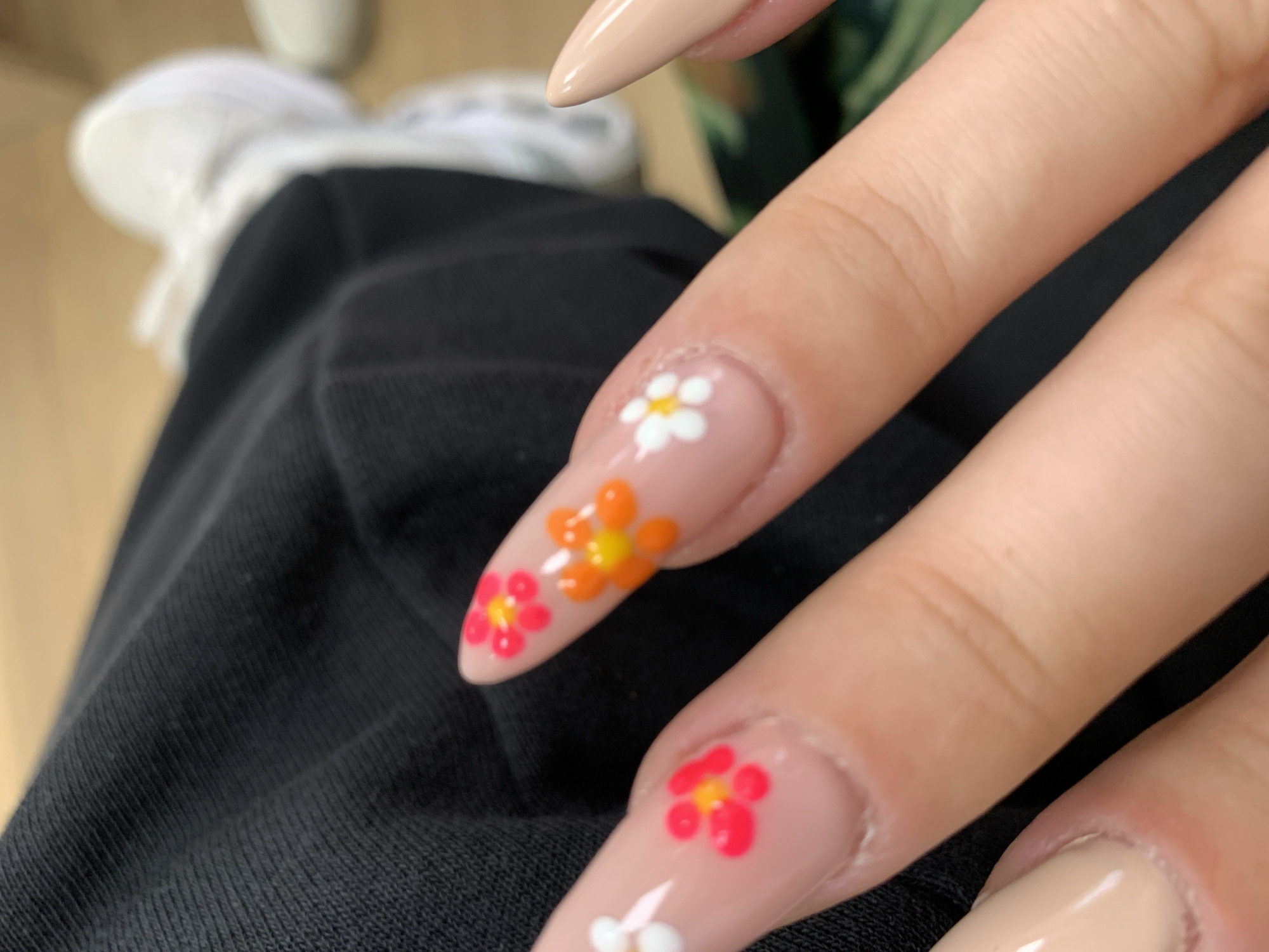 The image displays a close-up view of a person's hand with manicured fingernails. The nails are long and pointed (stiletto-shaped) with a nude or beige base color. Each nail is adorned with a floral design. One nail features a white flower, another has orange flowers, and a third has red flowers. The hand is resting on what appears to be a black garment, possibly clothing. In the background, a white shoe is partially visible, suggesting the person is wearing it. The context of the image does not directly give the location as Sofia, Bulgaria.