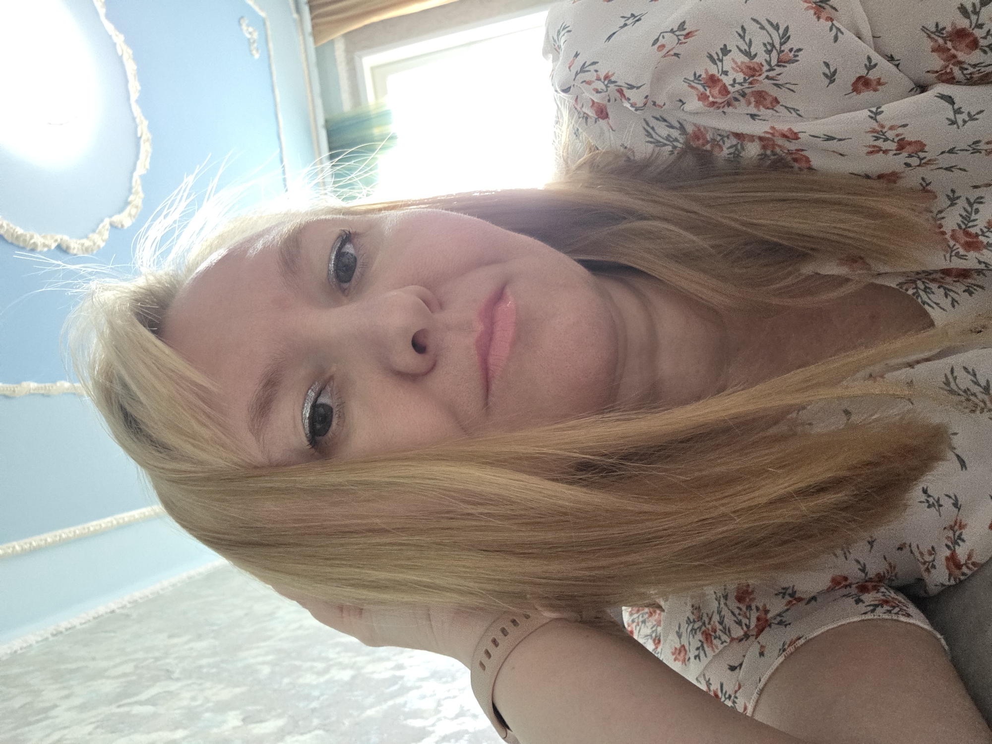 This is a slightly tilted, close-up, low-angle selfie of a woman with blonde hair. She is wearing a white dress with a floral pattern of red and green flowers. Her left eye, which is visible, has glittery eyeshadow. Her lips are painted with a light pink lipstick and appear slightly glossy. Her right hand is supporting her head, and she is wearing a light pink smartwatch on her left wrist. The background consists of a light blue wall with decorative molding, and a window with natural light streaming through. The overall lighting suggests it's daytime. There are no visible signs of text in the image. The scene appears to be indoors, possibly a bedroom or living room, in Novocheboksarsk, Russia. The woman's expression is neutral.